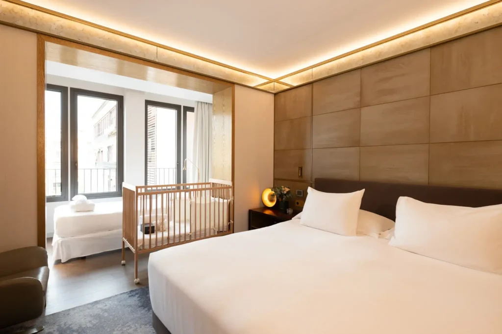Modernes Hotelzimmer mit einem großen Bett, einem Babybett, einem Einzelbett am Fenster, einem Sessel und neutralem Dekor. Natürliches Licht fällt durch die bodentiefen Fenster ein.