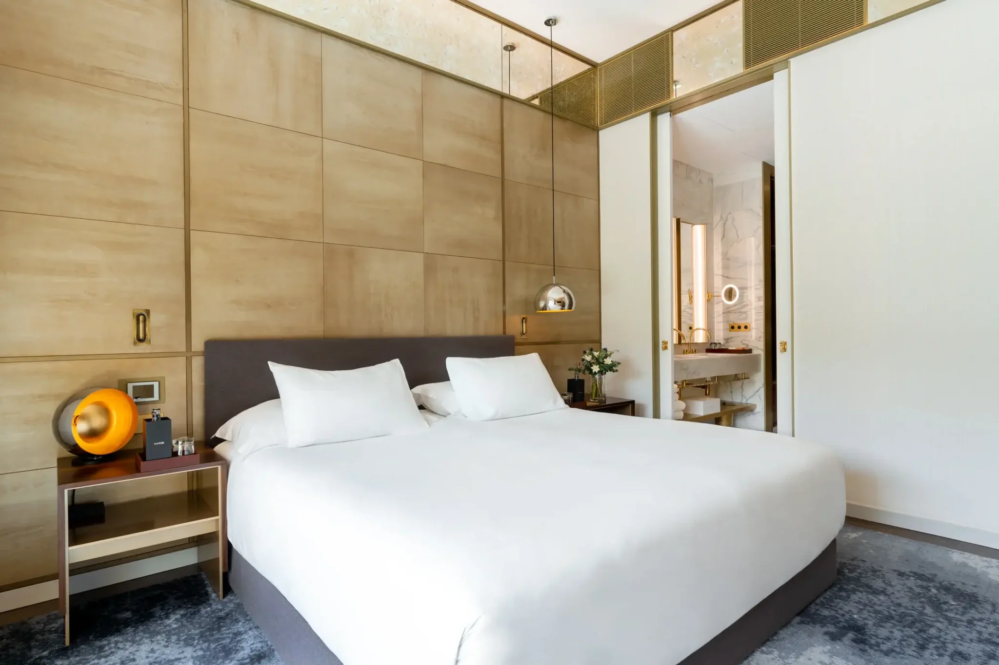 Dormitorio moderno con cama grande, ropa de cama blanca, dos mesillas de noche y una puerta abierta que da a un cuarto de baño con paredes de mármol y detalles dorados.
