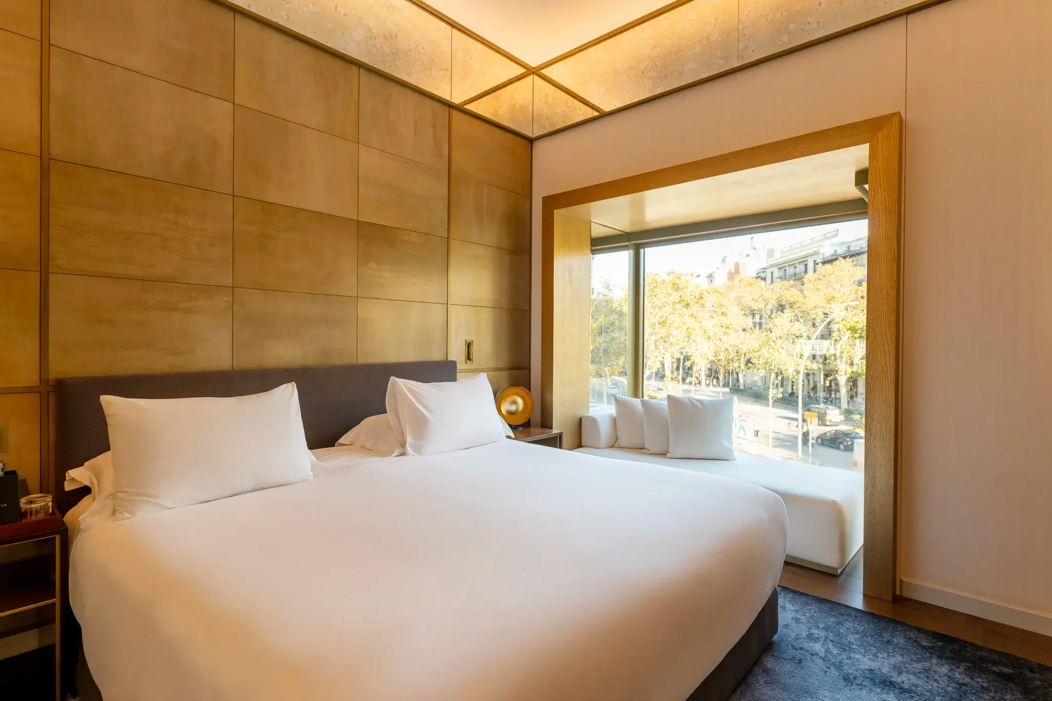 La moderna habitación Almanaque cuenta con una gran cama con sábanas blancas, paredes con paneles de color tostado y una zona de estar con ventana con vistas a una calle arbolada.