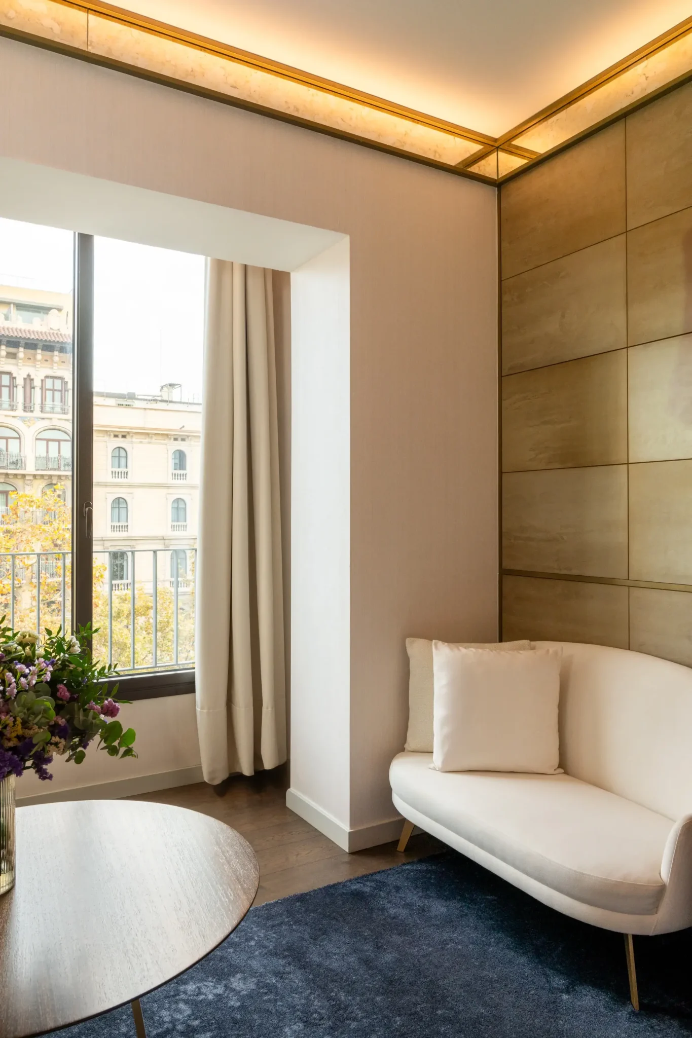 Una Habitació Almanac moderna amb un sofà de dues places encoixinat blanc, una taula rodona de fusta amb flors, una gran finestra amb cortines que donen al balcó i panells de paret en tons daurats.