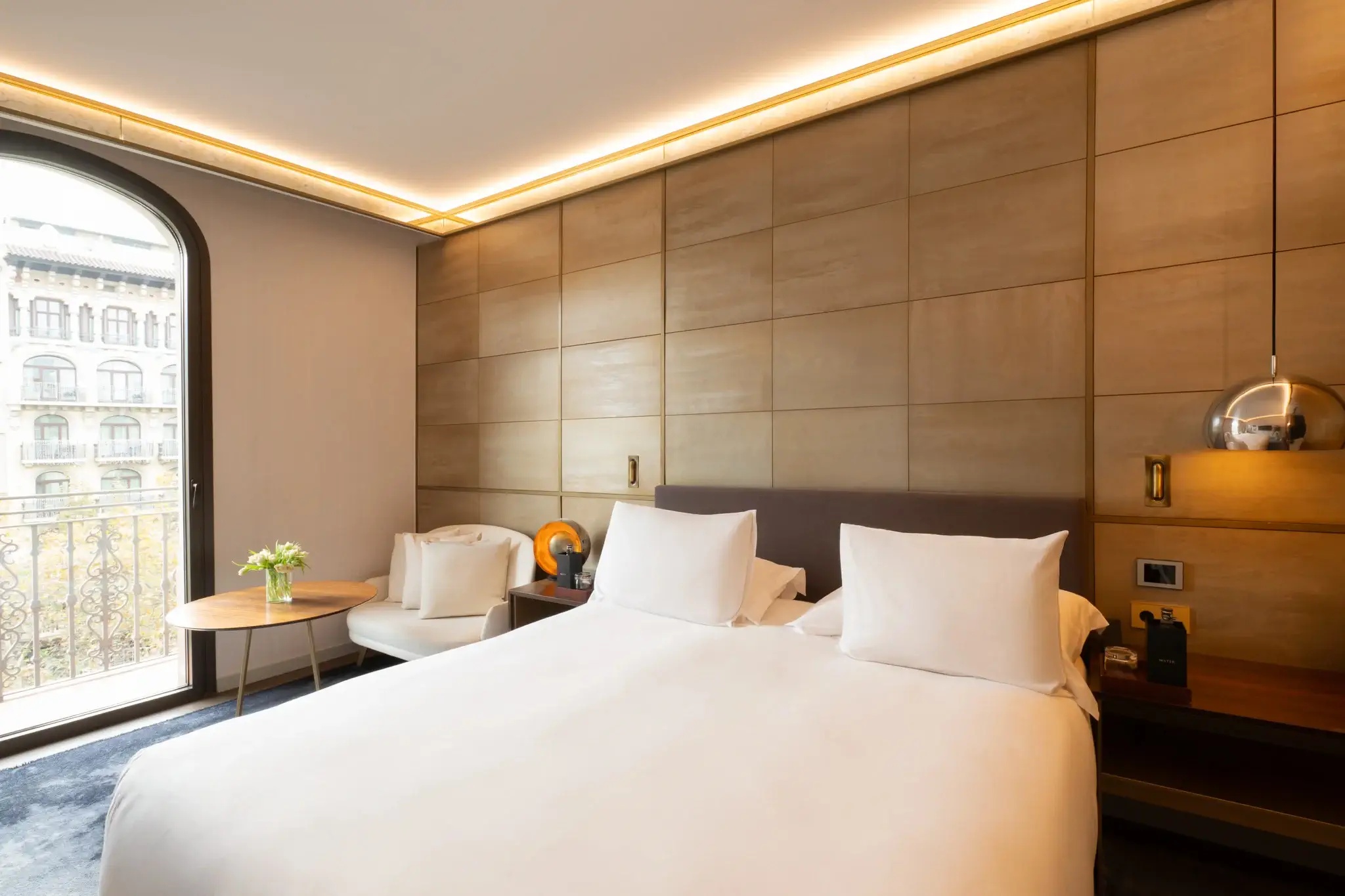 Habitació d'hotel moderna amb un llit gran, roba de llit blanca, tauletes de nit i una taula rodona amb flors. L'Habitació Almanac disposa d'una butaca i una gran finestra que deixa entrar llum natural i que dóna a un balcó privat.