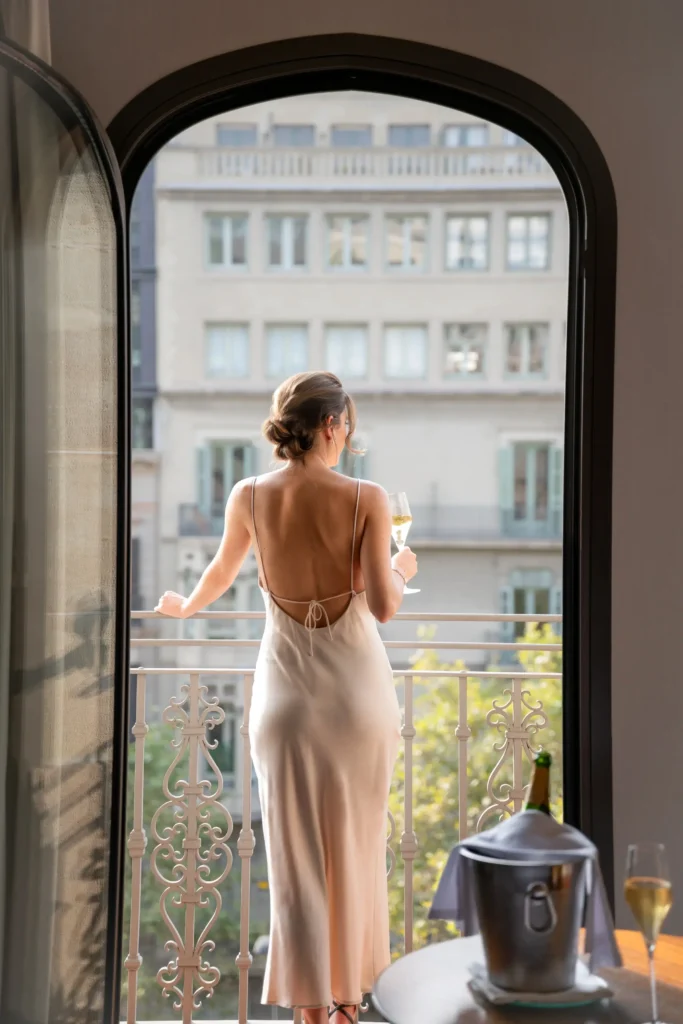 Una dona amb un vestit sense esquena es troba al balcó de l'Almanac de l'Habitació, amb una copa a la mà i contemplant els edificis de la ciutat a través d'una porta oberta.