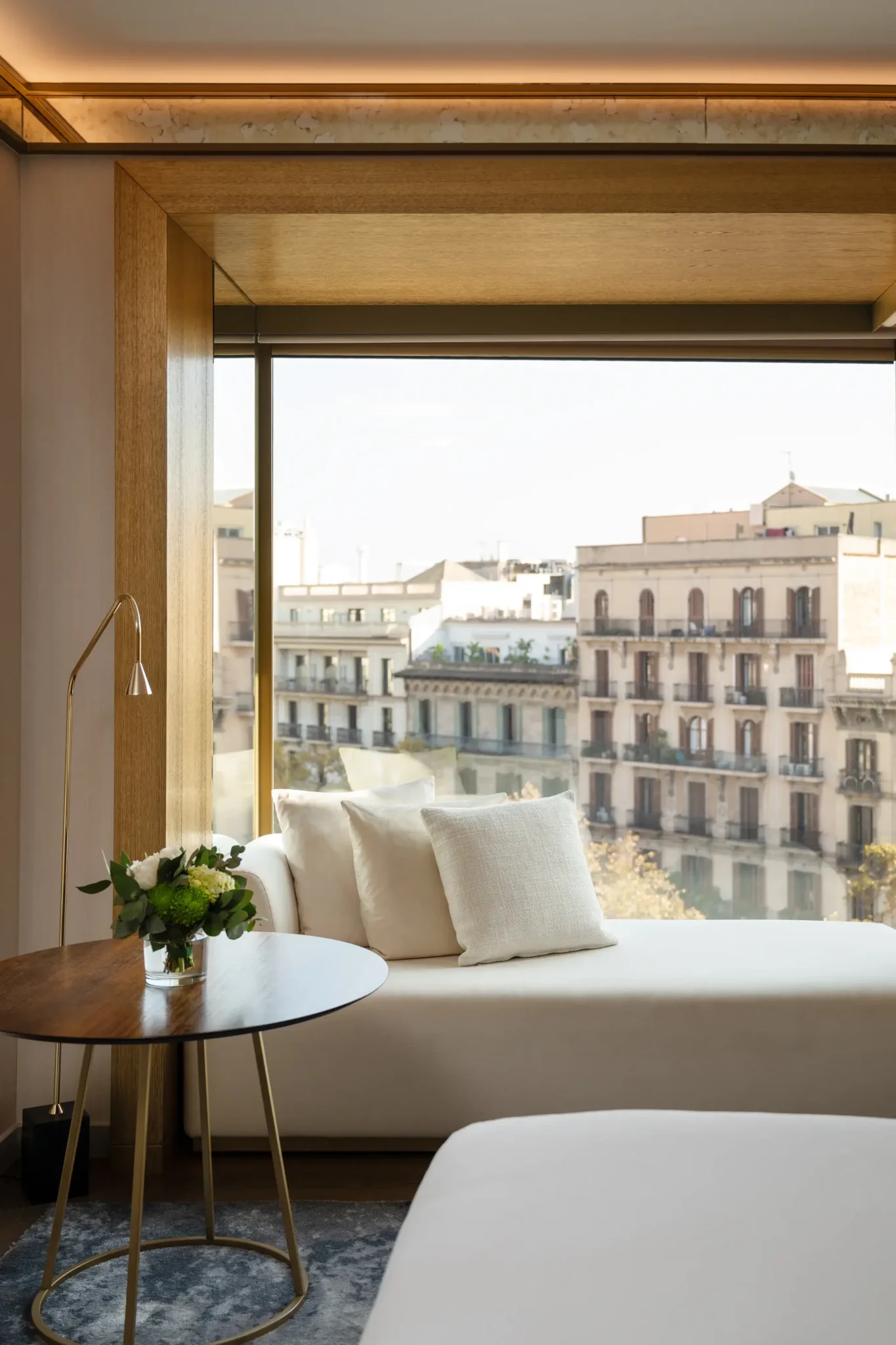 Disfrute de la Habitación Almanaque, una moderna habitación de hotel con un asiento en la ventana, dos almohadas blancas, una pequeña mesa redonda con un arreglo floral y unas impresionantes vistas de los edificios de la ciudad.