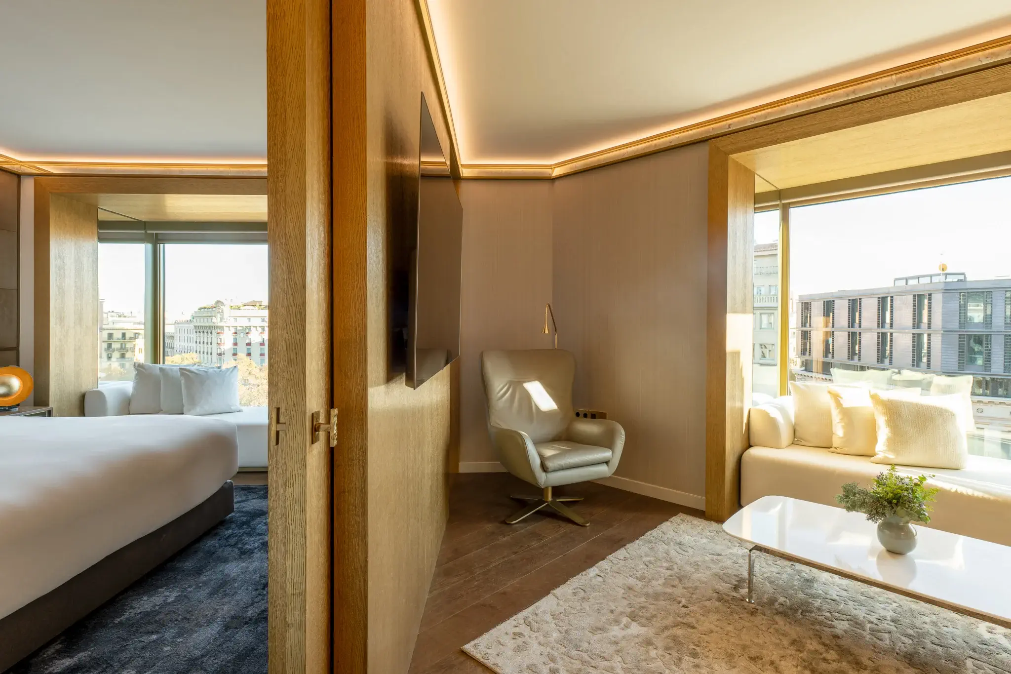 Modern Almanac Suite with a bedroom and adjoining sitting area, featuring large windows, a white armchair, a sofa, and views of city buildings outside.