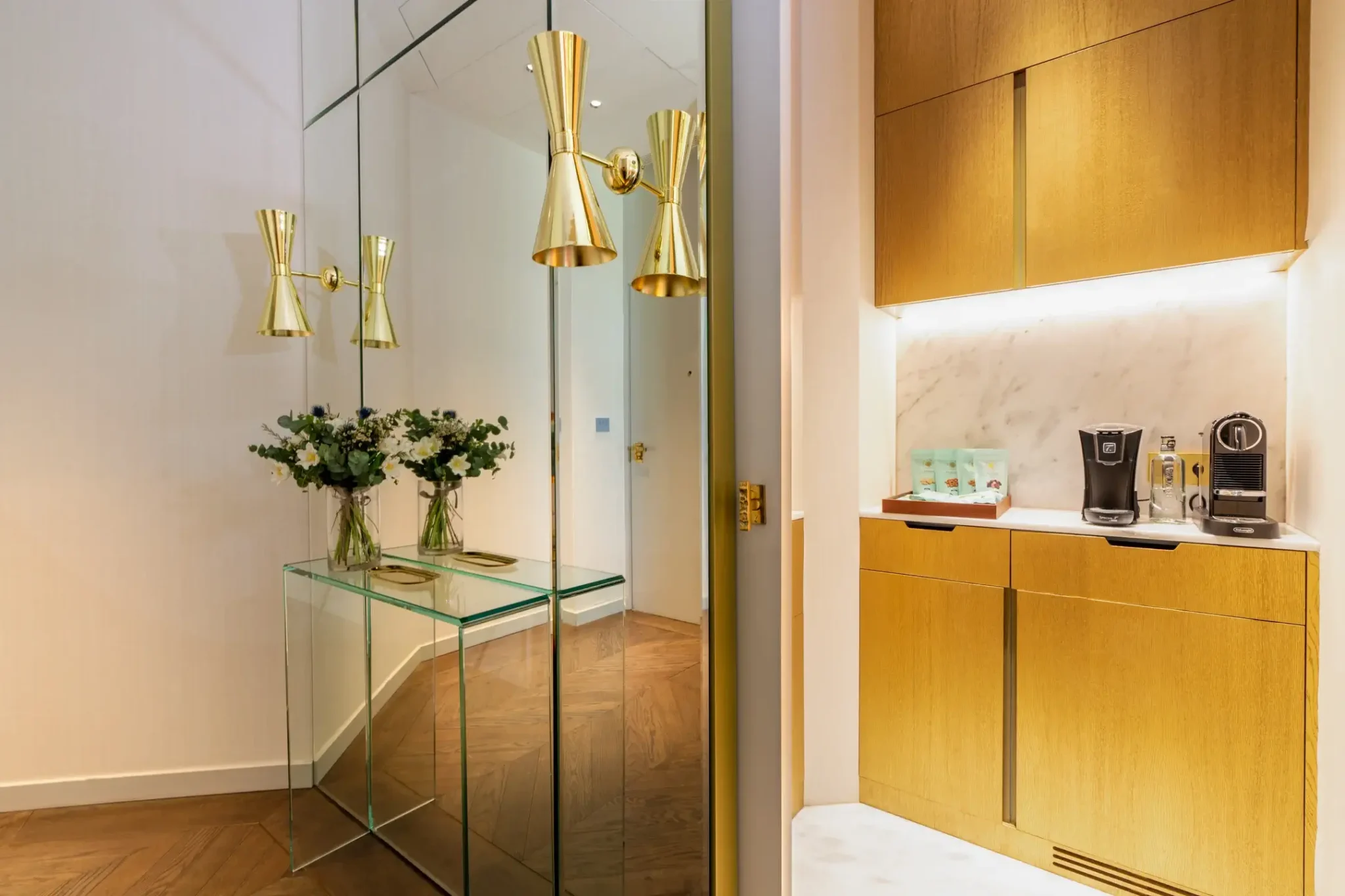 A modern entryway in the Almanac Suite features mirrored walls, gold sconces, two vases of flowers on a glass console, and an adjacent coffee station with gold cabinets and sleek coffee machines.