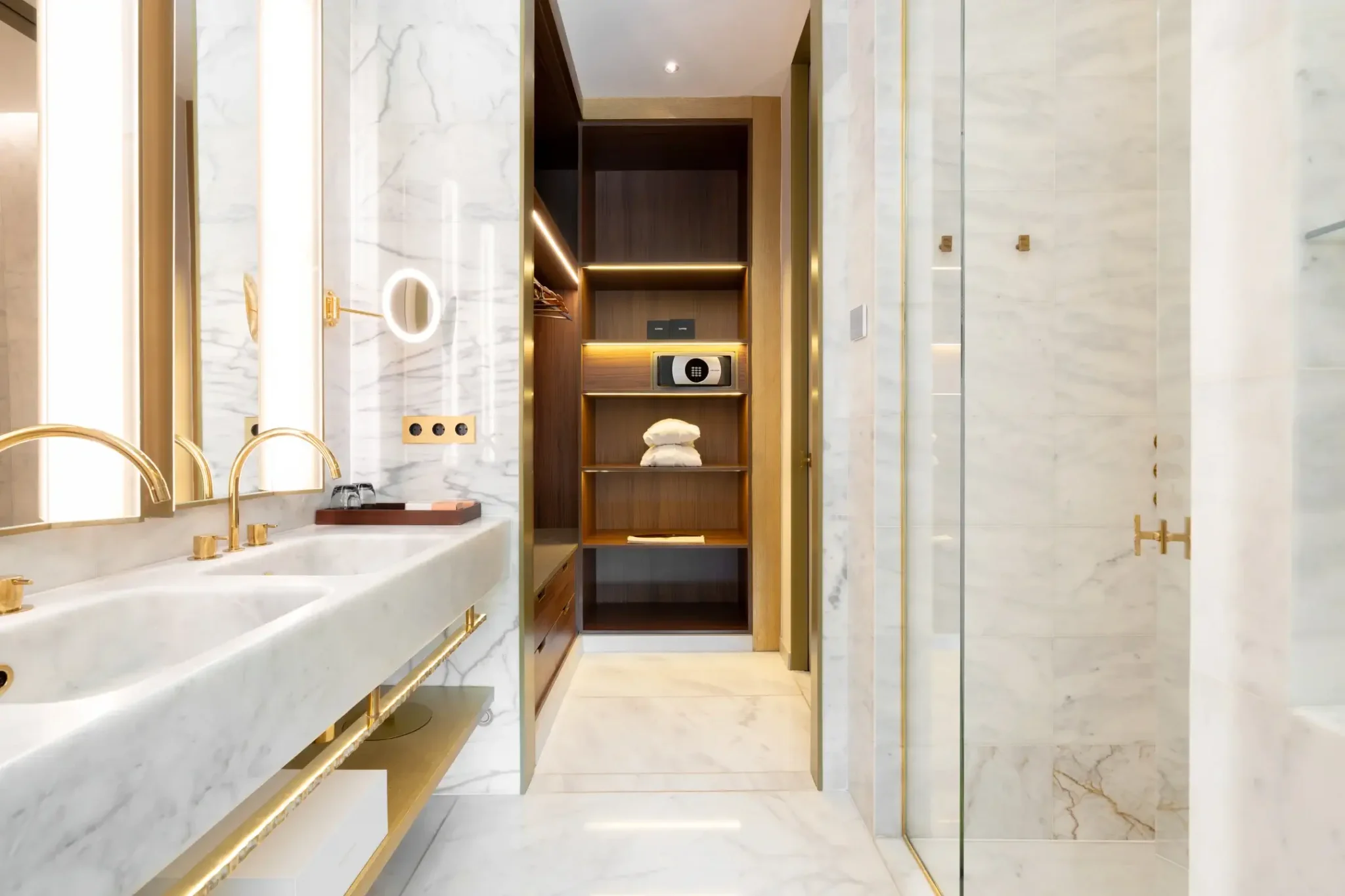 Modern Almanac Suite bathroom with marble surfaces, double sinks, gold fixtures, a glass shower, and an open closet with shelves for towels and a safe.