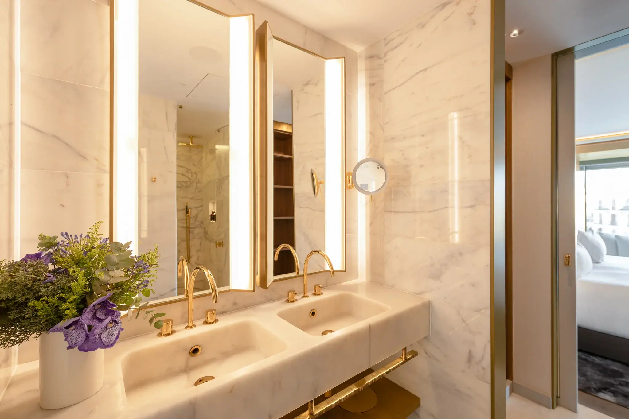 Modern bathroom in the Almanac Suite features marble walls, double sinks with gold fixtures, large mirrors with integrated lighting, a bouquet of flowers, and a partial view of the bedroom through an open door.