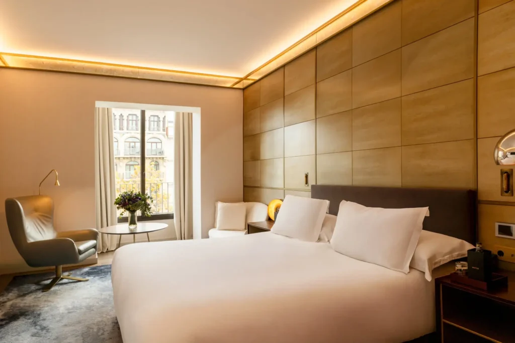 Ein modernes Hotelzimmer mit einem großen Bett, zwei weißen Stühlen am Fenster, einem kleinen runden Tisch mit Blumen und warmer Beleuchtung an hellbraun getäfelten Wänden.