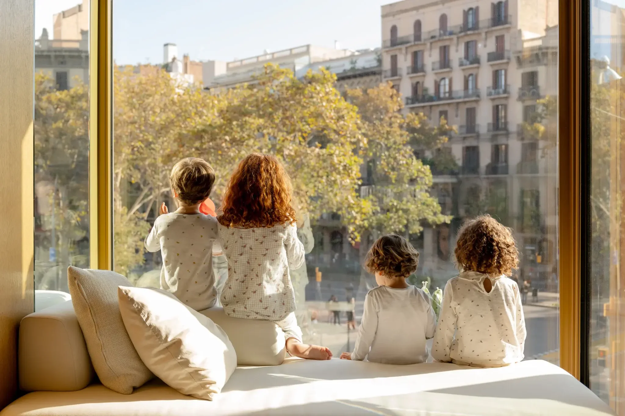 Vier Kinder in Schlafanzügen sitzen auf einer Fensterbank und blicken an einem sonnigen Tag auf die Gebäude und Bäume der Stadt.