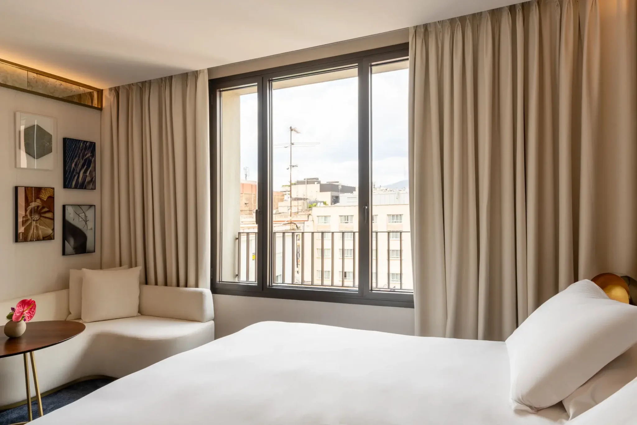 Modernes Hotelzimmer mit einem großen Bett, weißer Bettwäsche, einem kleinen runden Tisch mit einer Blumenvase, einem gepolsterten Stuhl und einem Fenster mit Stadtblick und beigen Vorhängen.