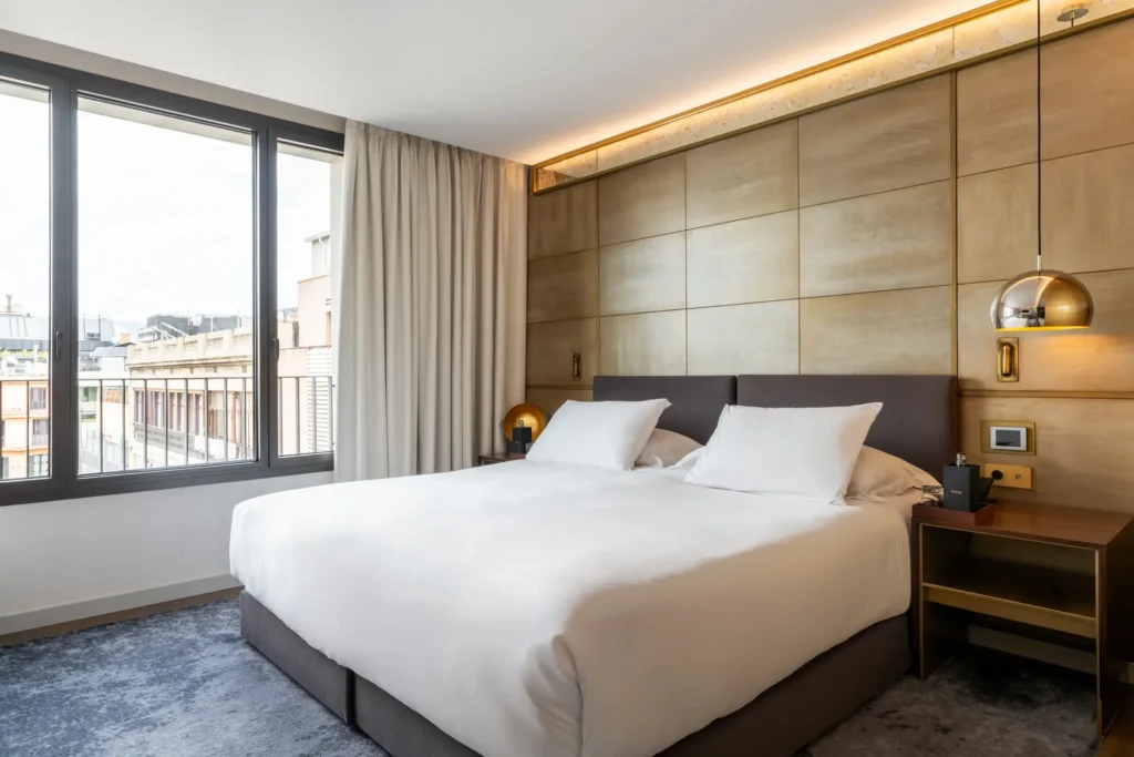 Ein modernes Hotelzimmer mit einem großen Bett, zwei Kissen, Nachttischen mit Lampen, bodentiefen Fenstern, beigen Wandpaneelen und grauem Teppich.