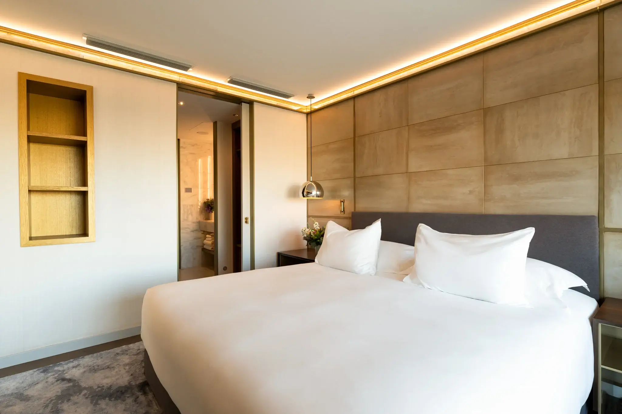 Moderne Almanac Suite mit großem Bett, weißer Bettwäsche, gepolsterter beiger Akzentwand, eingebauten Regalen, Nachttischen und offener Badezimmertür im Hintergrund - perfekt für Gäste, die zwei Schlafzimmer benötigen.