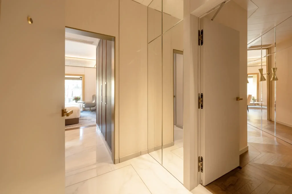 Ein Flur mit beigen Wänden, verspiegelten Paneelen, Marmorfußboden und Türen, die zu hellen Zimmern führen, wie der eleganten Almanac Suite und zwei Schlafzimmern, die alle modern eingerichtet sind.