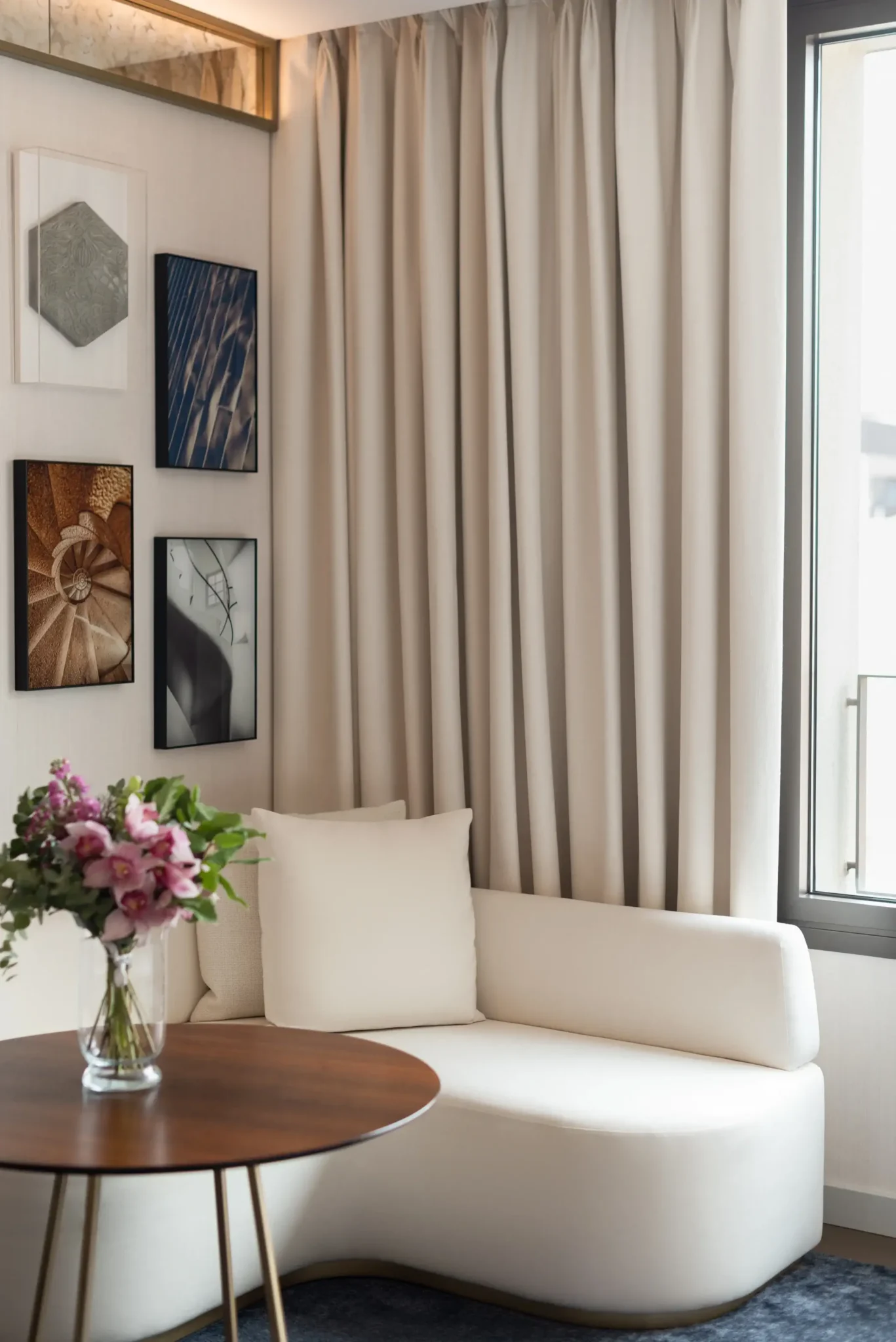 Una pequeña y moderna Sala de Galería con una tumbona blanca, una mesa redonda de madera con un jarrón de flores, cortinas beige, una ventana y cuadros en la pared.