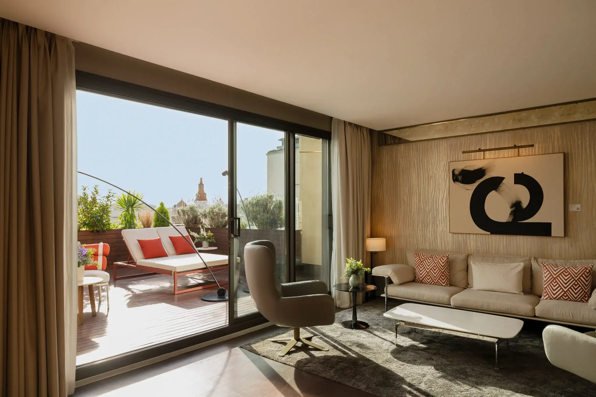 Das moderne Wohnzimmer der Grand Penthouse Suite verfügt über ein Sofa, einen Sessel, abstrakte Wandkunst und Glasschiebetüren, die zu einer sonnenbeschienenen Terrasse mit Sitzgelegenheiten im Freien und Topfpflanzen führen.