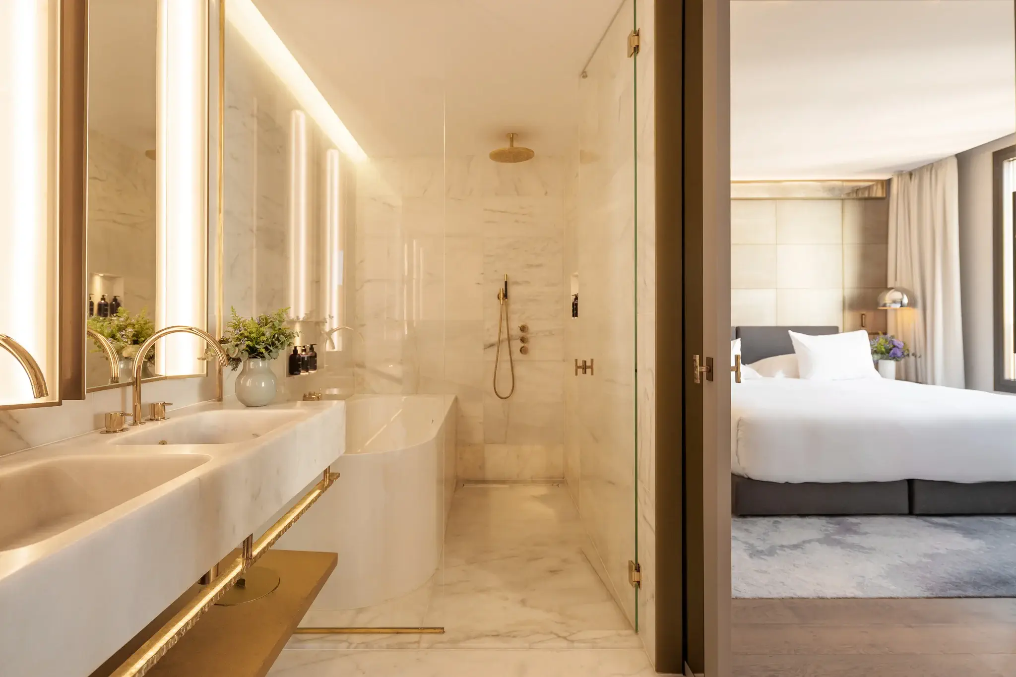 Modern hotel bathroom in the Grand Penthouse Suite features double sinks, bathtub, rain shower, and gold fixtures, adjacent to a bedroom with a neatly made bed and large window.
