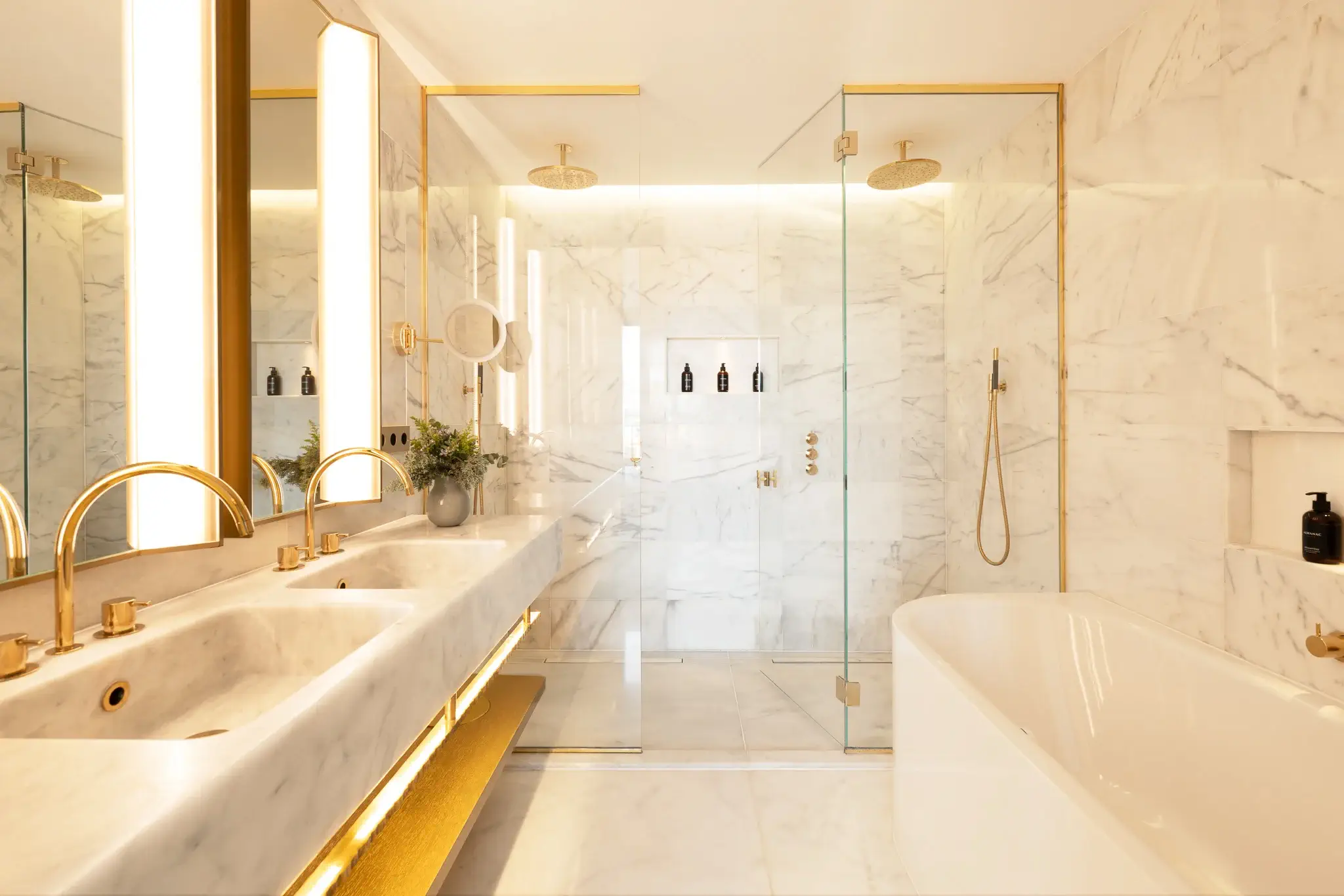 Modern Grand Penthouse Suite bathroom with double sinks, gold fixtures, a walk-in glass shower, marble walls, built-in shelves, and a white bathtub.
