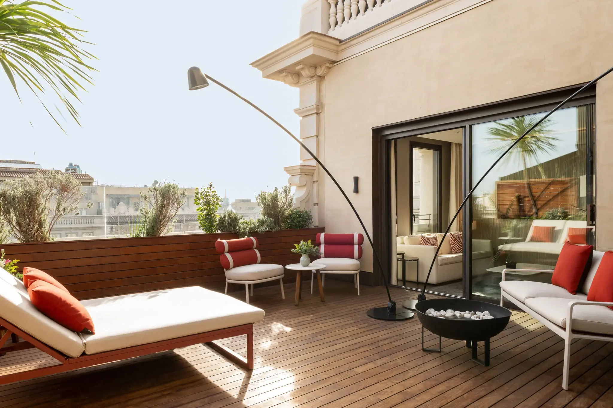 A modern outdoor patio in the Grand Penthouse Suite features wooden flooring, lounge chairs with red and white cushions, a sleek black fire pit, and potted plants near the building.