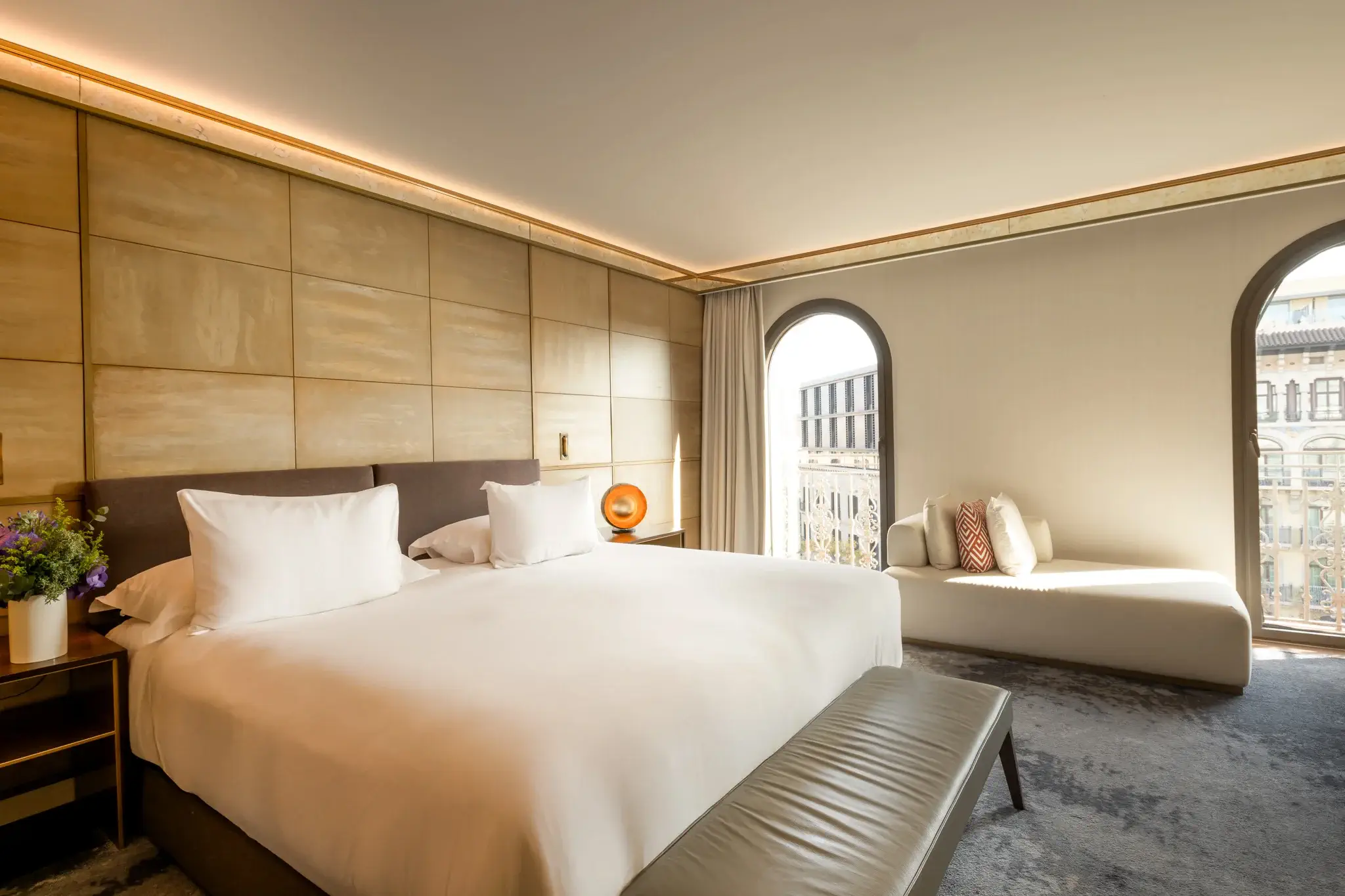 Experimenta la nostra Junior Suite: una habitació d'hotel moderna amb un llit gran, un banc gris, una tauleta auxiliar amb flors i una butaca al costat de dues finestres arquejades que omplen l'espai de llum natural.