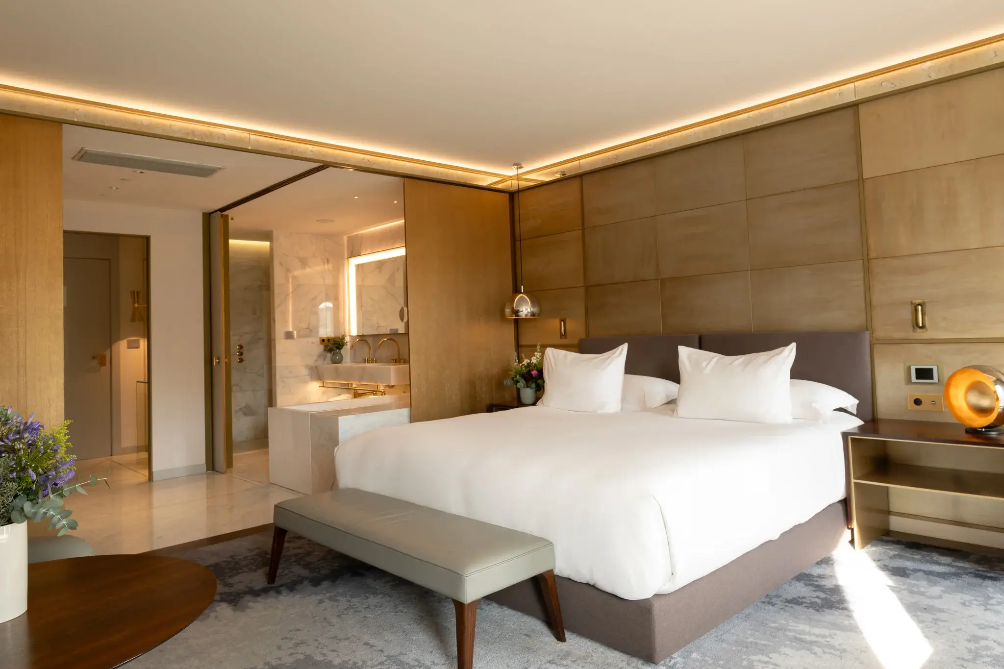 Experimenta una espaiosa Junior Suite amb un llit king size, roba de llit blanca, capçal encoixinat, banc, tauleta de nit i vistes obertes a un bany de marbre amb una banyera de luxe.