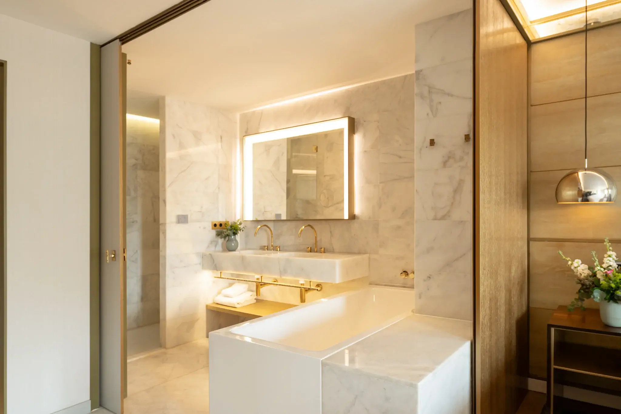 Un bany modern de tipus Junior Suite amb parets de marbre, un gran mirall amb retroiluminació, un lavabo doble, una banyera integrada i un gerro de flors sobre una tauleta auxiliar.