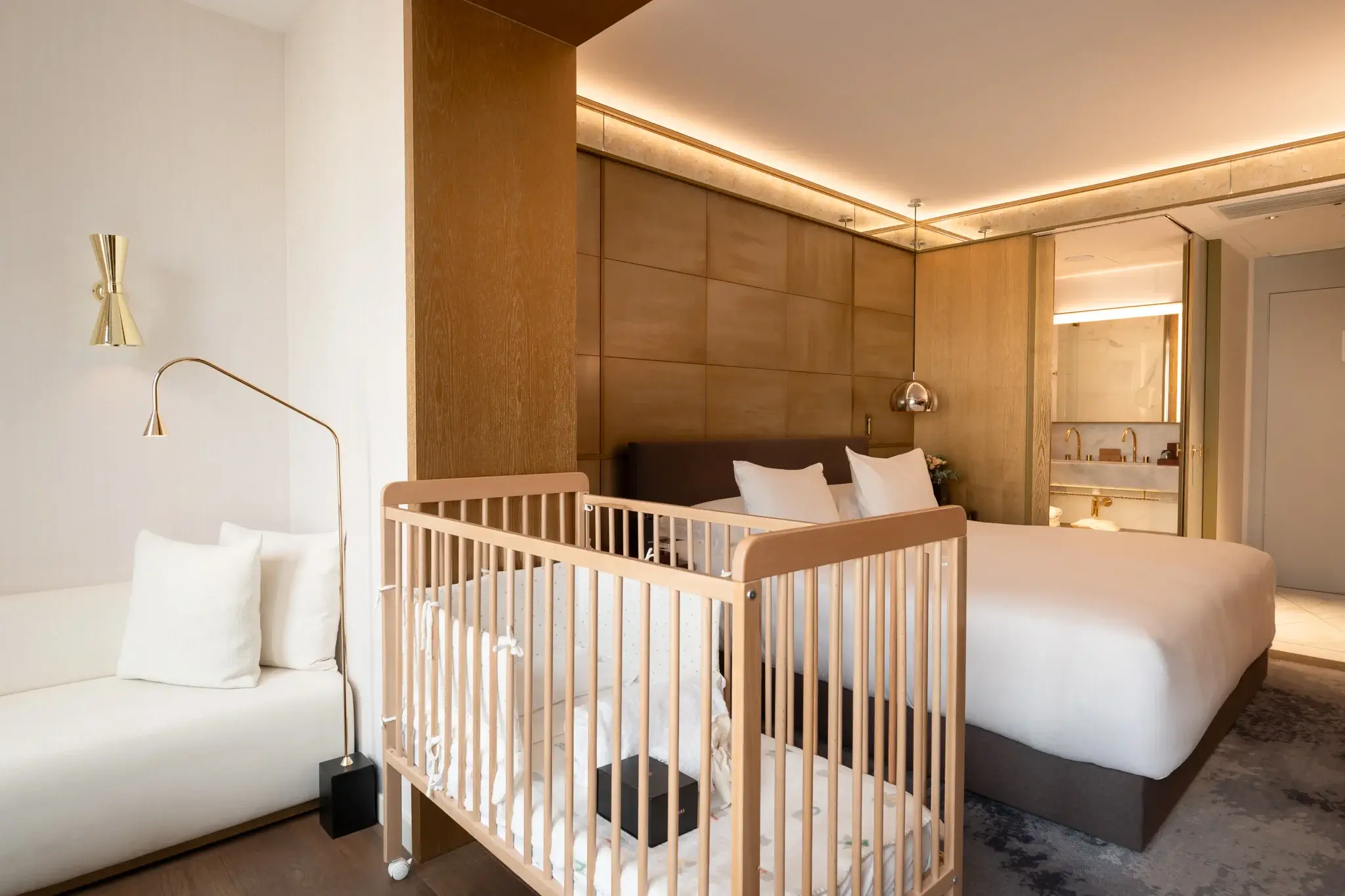Una habitación de hotel con una cama doble, una cuna de madera, un sofá blanco y vistas a la zona del cuarto de baño con lavabo y espejo.