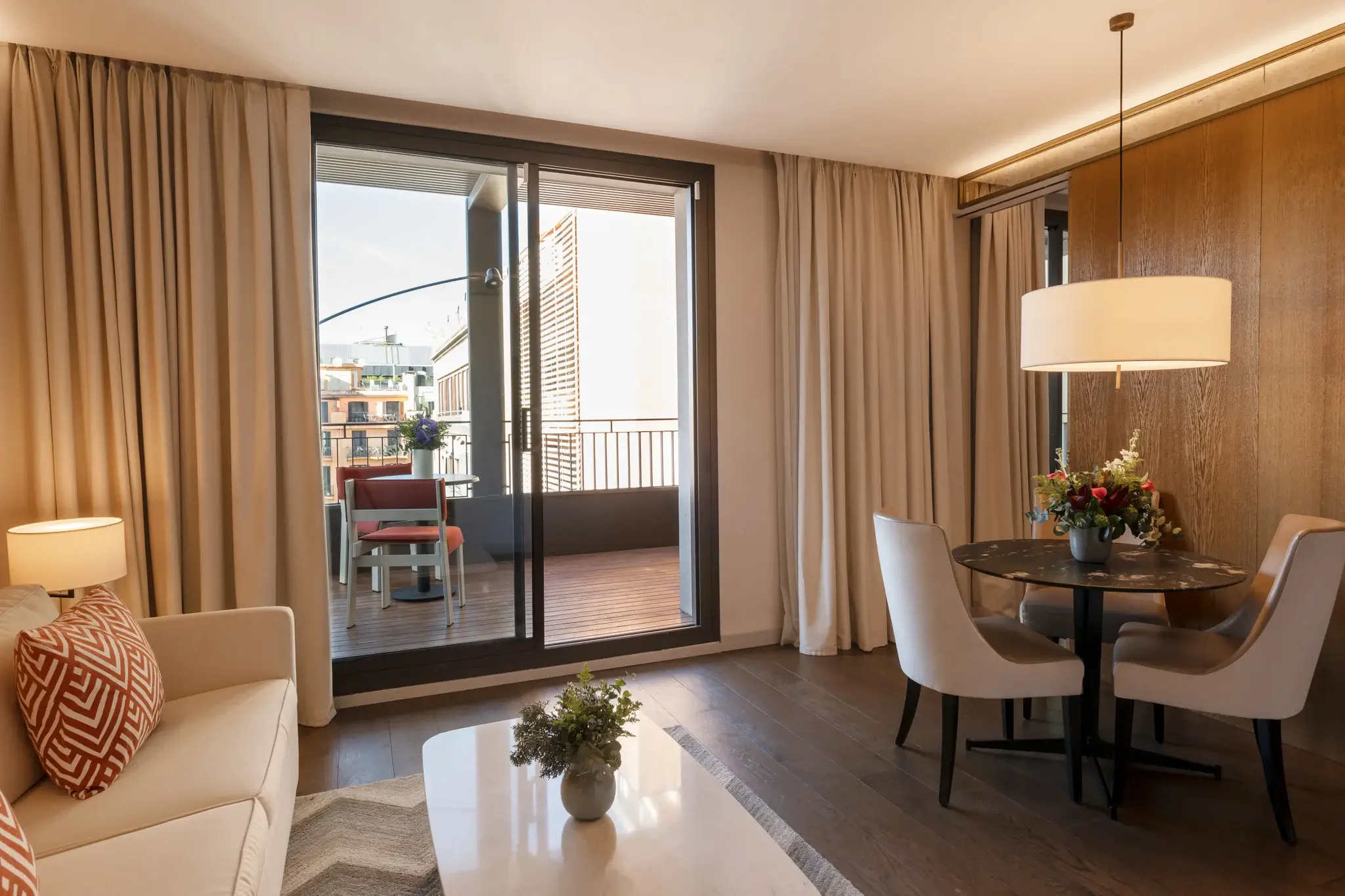 A modern Terrace Suite living room with a sofa, coffee table, small dining area, and large sliding glass doors leading to a balcony with outdoor seating.