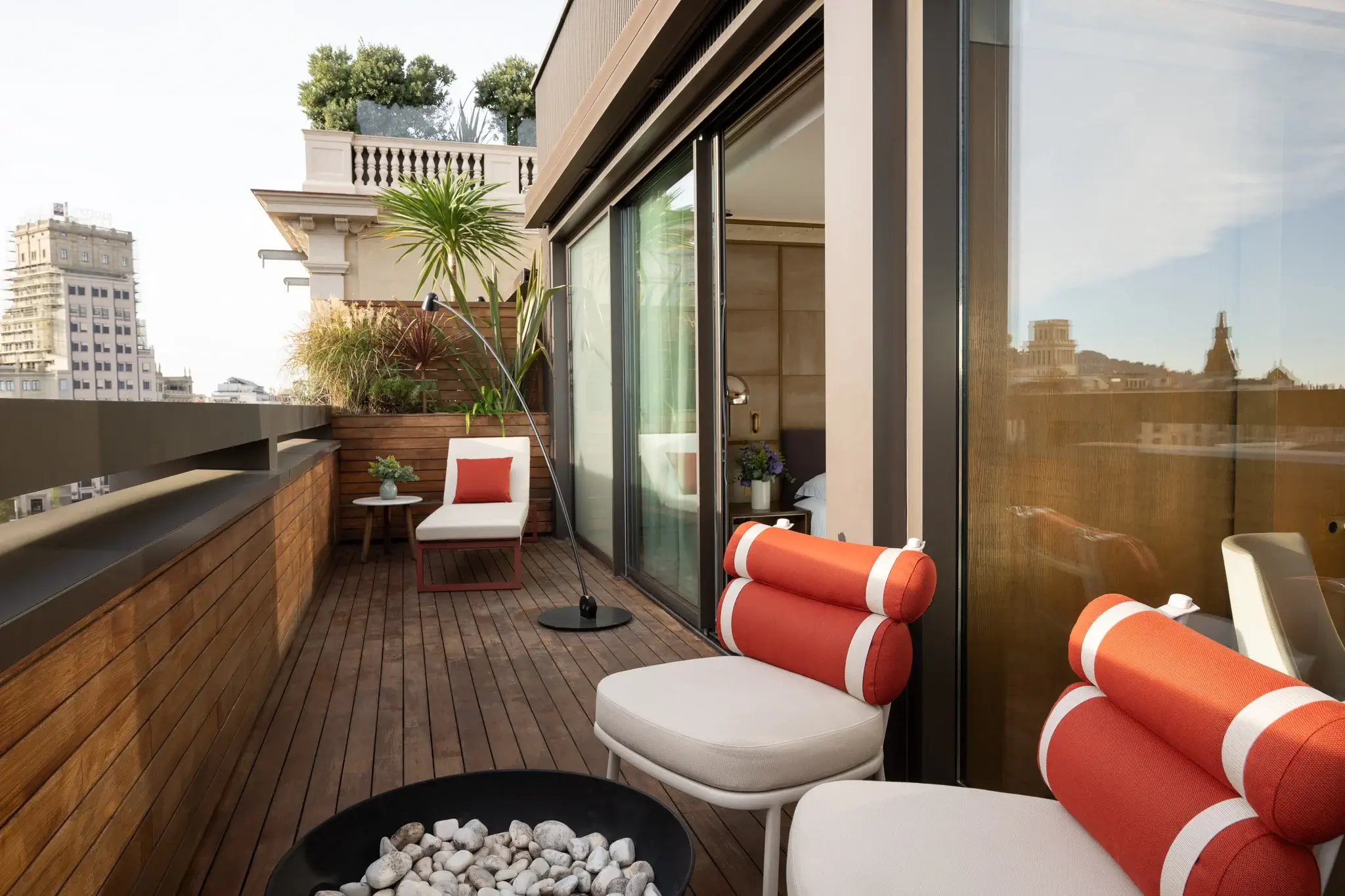 A modern Terrace Suite balcony with wooden flooring, white and orange cushioned chairs, a small lounge area, potted plants, and a stunning cityscape view in the background.