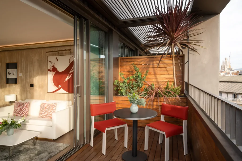 A modern Terrace Suite balcony with a round table, two red chairs, potted plants, and sliding glass doors opening to a living room featuring a white sofa and abstract art.