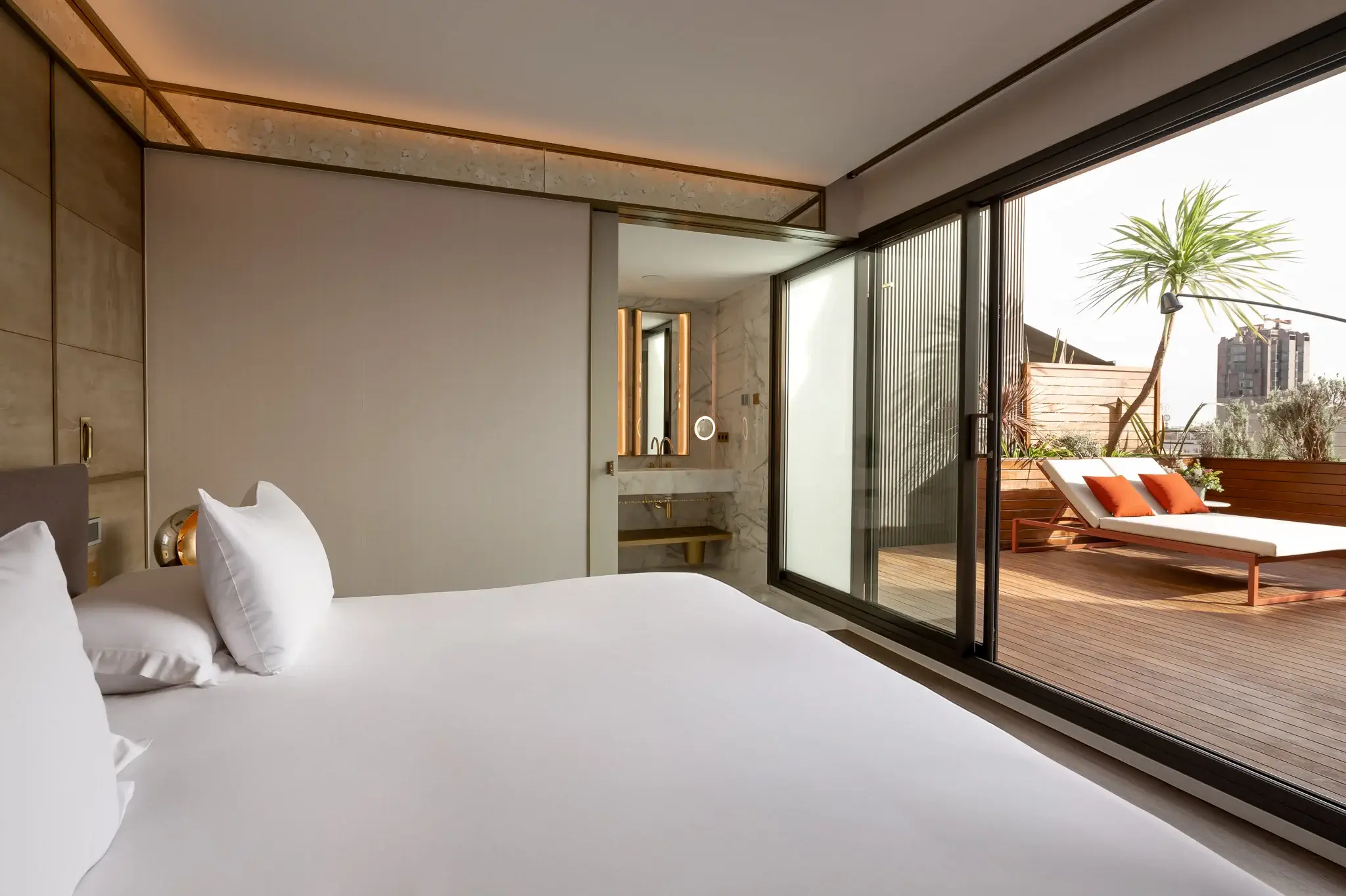 Modern Terrace Suite featuring a neatly made bed, sliding glass doors opening to a private wooden terrace with lounge chairs and a potted palm, plus an ensuite bathroom in the background.