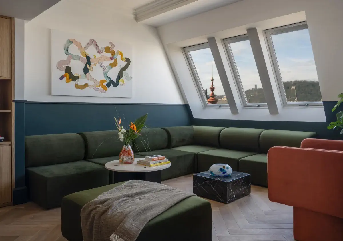 Modernes Wohnzimmer mit grünem Sektionssofa, orangefarbenem Sessel, abstrakter Wandmalerei und großen Fenstern, die an die Prager Hotels Almanac und Alcron erinnern, sowie einem Marmorcouchtisch und einem kleinen runden Tisch mit Blumen.