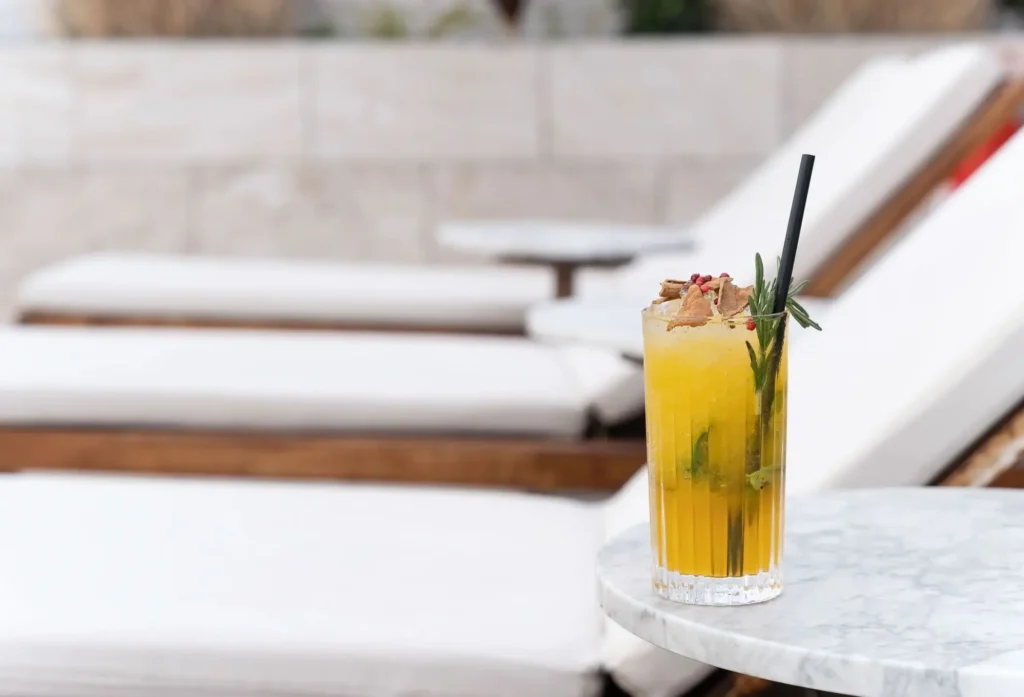 A yellow cocktail with herbs and ice sits on a round marble table beside white lounge chairs at Azimuth Rooftop Bar.