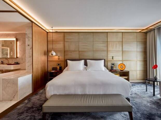 Moderne Junior Suite mit einem Doppelbett mit weißem Bettzeug, Nachttischen, einer Marmorbadewanne auf der linken Seite und großen Fenstern mit Vorhängen auf der rechten Seite.