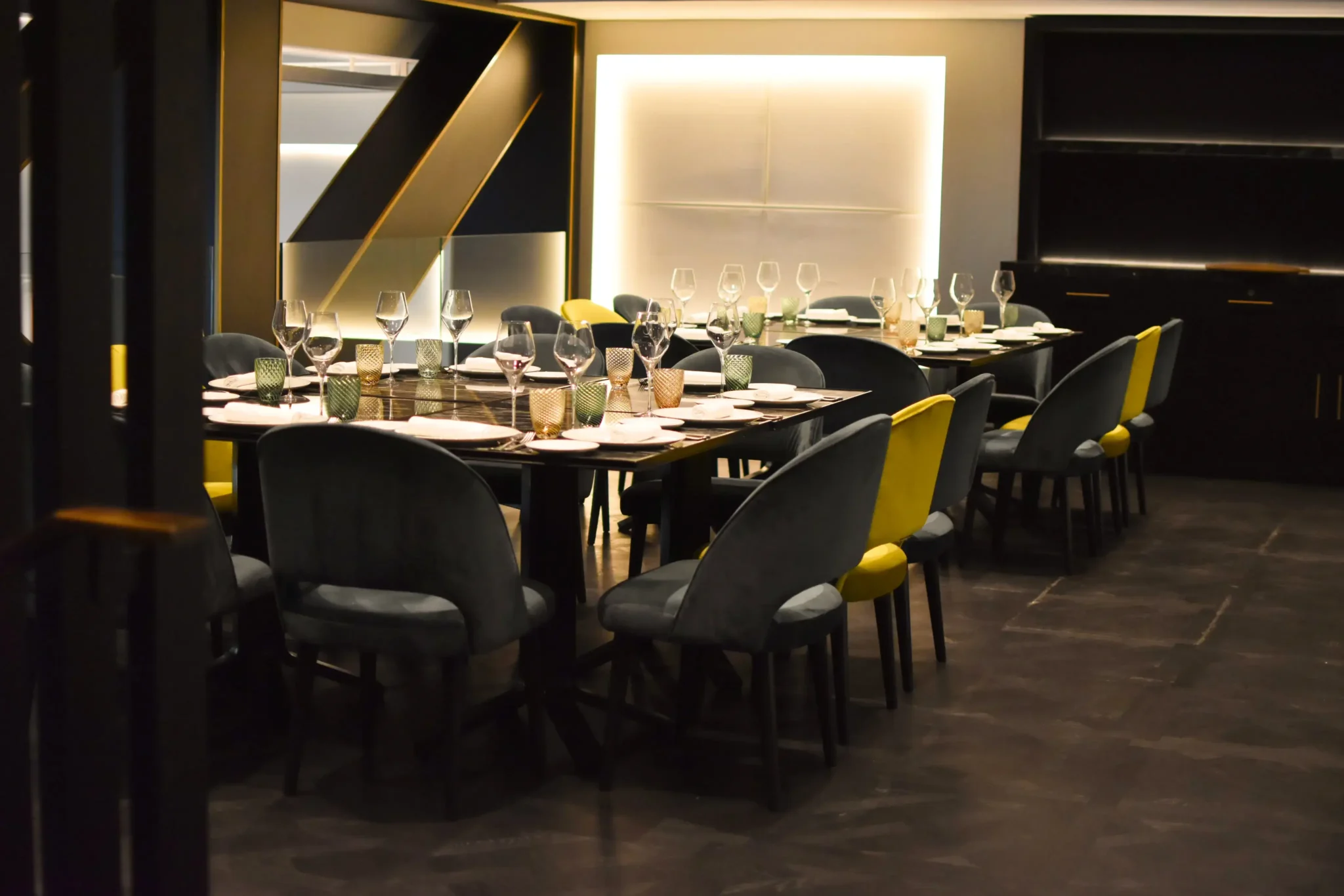 Un comedor moderno con dos mesas largas preparadas para eventos sociales, con platos, vasos y cubiertos. Las sillas son una mezcla de gris oscuro y amarillo, que complementa la decoración minimalista de la sala.