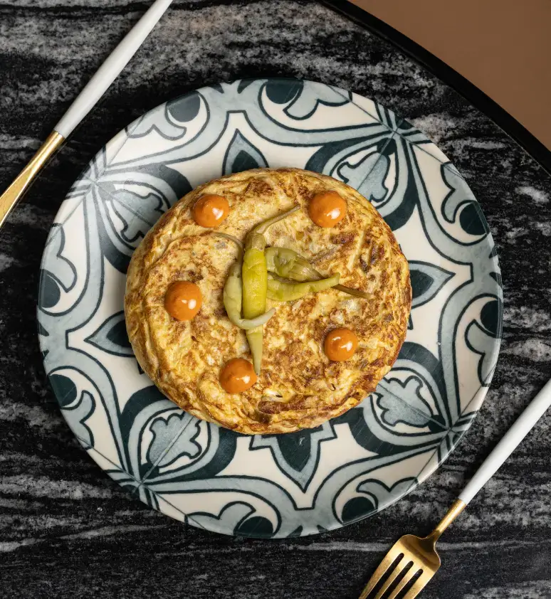 Tortilla española redonda con salsa y pimientos verdes cortados en rodajas, servida en un plato decorativo con cuchillo y tenedor dorados.