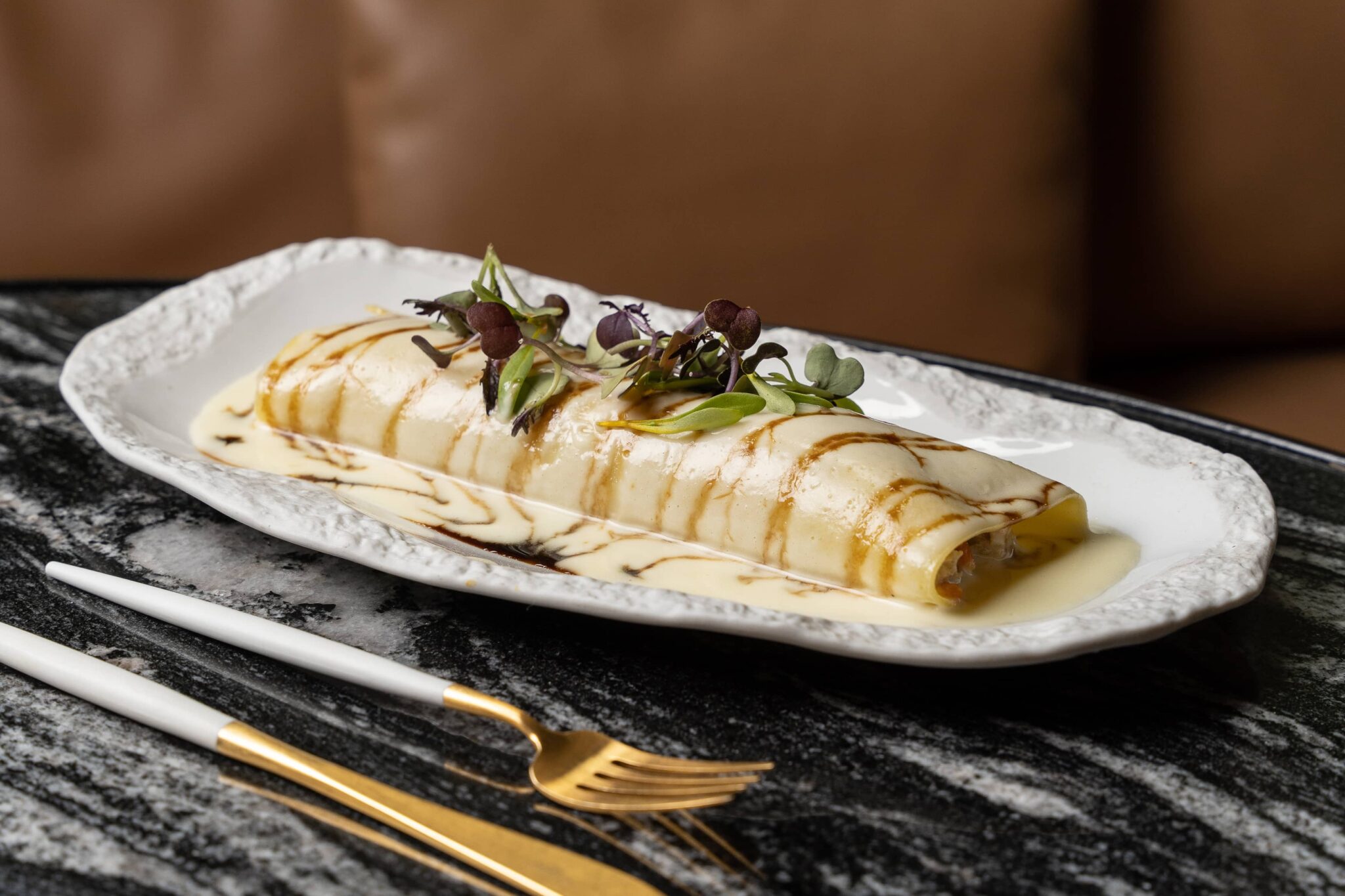Un crepe enrollado cubierto de microgreens y salsa, servido en un plato blanco con un tenedor y un cuchillo al lado sobre una mesa de mármol.