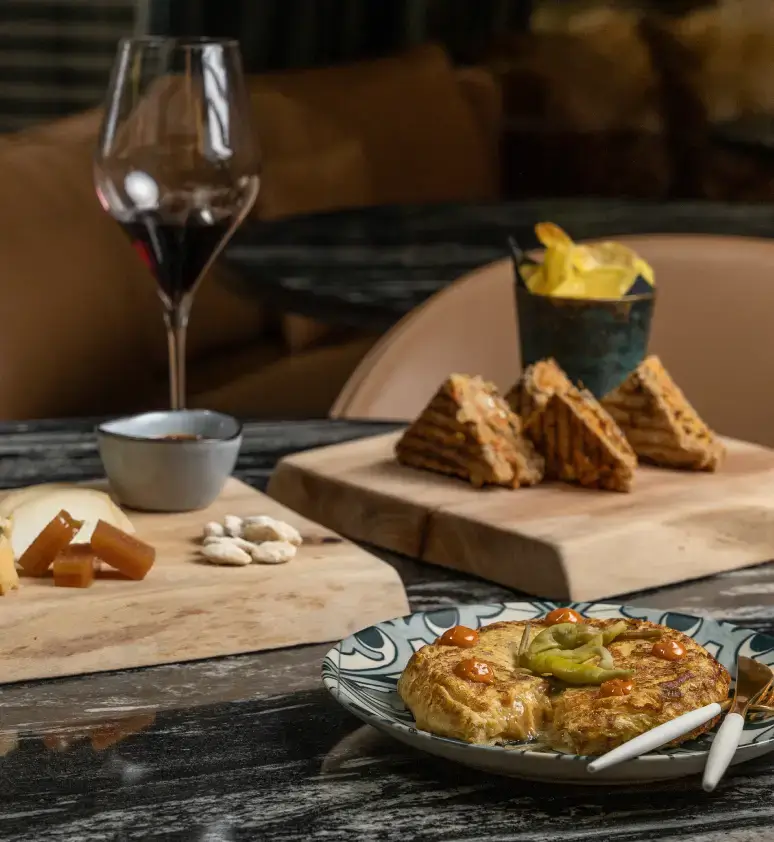 Una mesa puesta con un vaso de vino tinto, queso y frutos secos, bocadillos en una tabla de madera, patatas fritas y un plato de tortilla española adornada con pimientos.