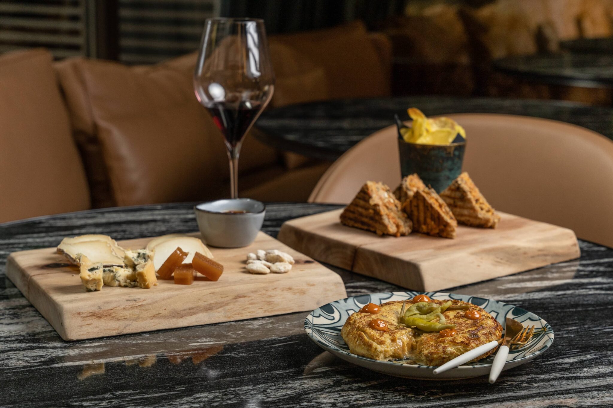 Una mesa con queso, pasta de fruta y galletas sobre tablas de madera, un plato de quiche con tenedor y cuchillo, un cuenco de patatas fritas y un vaso de vino tinto.