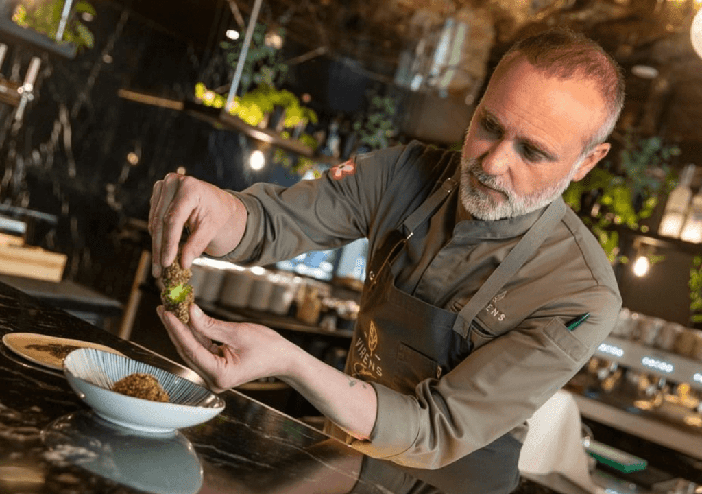 Un xef amb uniforme i davantal grisos serveix acuradament el menjar a la cuina moderna del restaurant Virens, amb plantes i estris de cuina visibles al fons.
