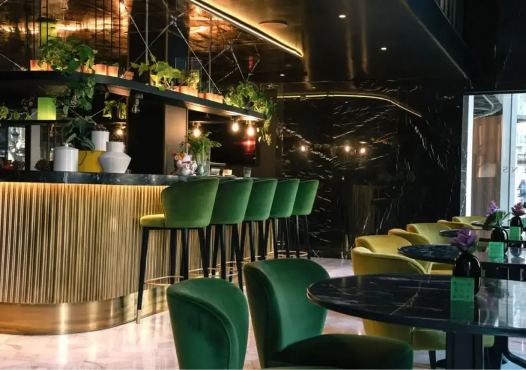 Moderno interior de bar con sillas de terciopelo verde, mesas de mármol, plantas en macetas e iluminación cálida, con una combinación de colores oscuros y una decoración elegante.