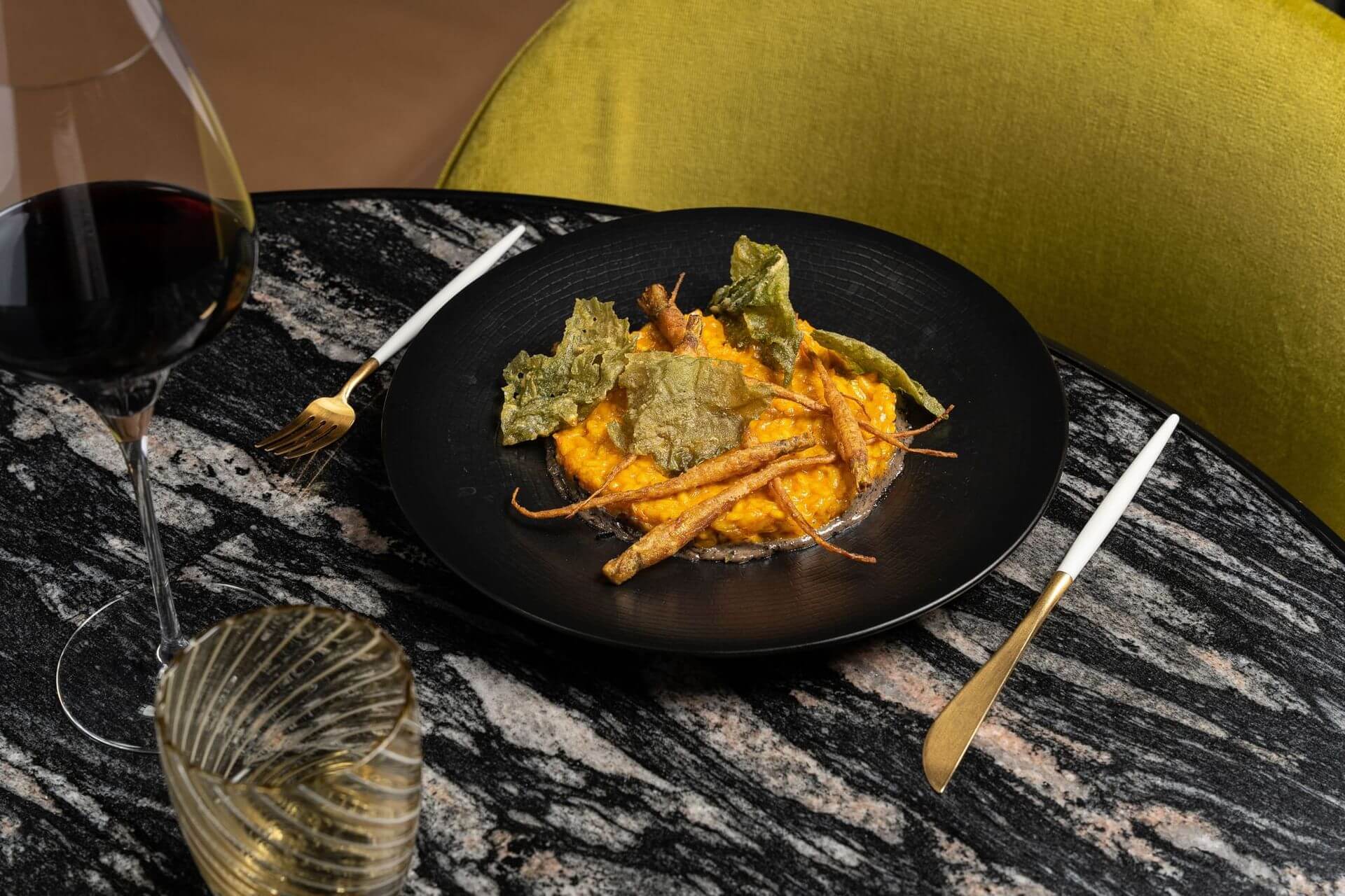 Un plat negre amb una ració de risotto cremós amb guarnicions de verdures cruixents es troba sobre una taula de marbre al costat d'una copa de vi negre i una copa d'aigua.