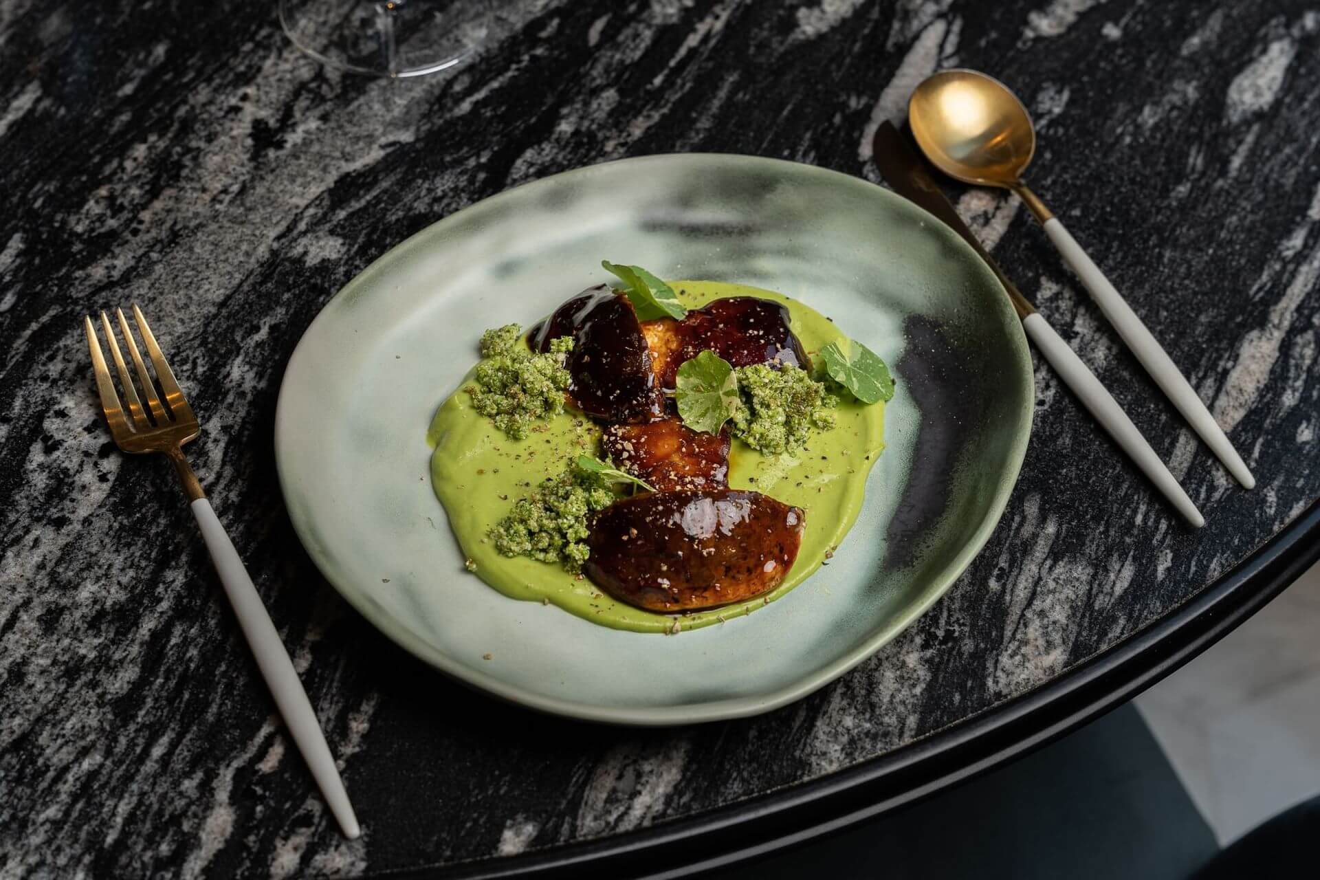 Un plat de ceràmica amb tres trossos de menjar glaçats sobre una salsa verda, guarnit amb crumble verd i microverds, està col·locat sobre una taula de marbre fosc amb coberts daurats i blancs.