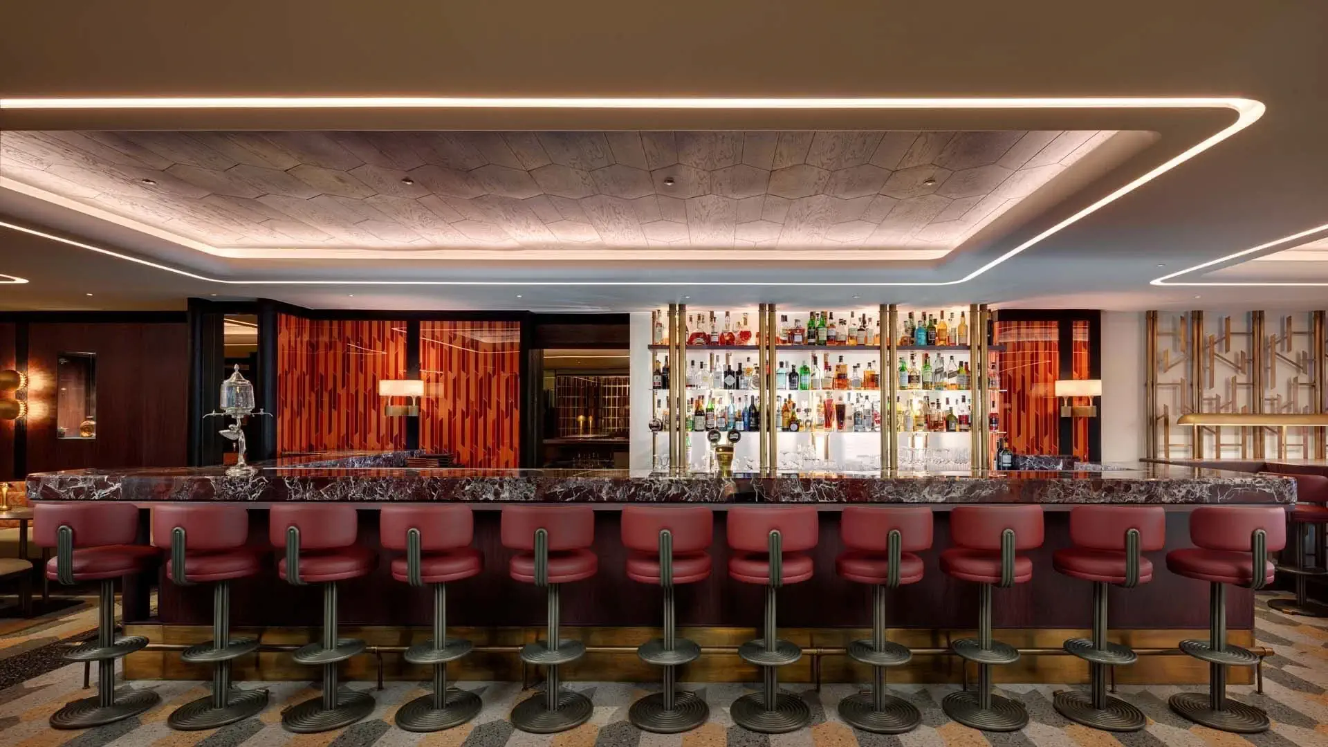 Eine moderne Bar mit Marmortheke, roten Barhockern und einer hinterleuchteten Auslage mit verschiedenen Flaschen in Glasregalen - ein einladender Ort, um Essen und Trinken bei warmer, stimmungsvoller Beleuchtung zu genießen.