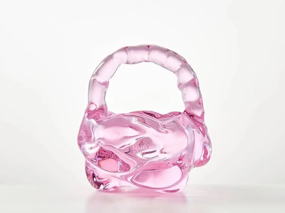 Diese skulpturale Handtasche aus rosafarbenem Glas mit abgerundetem Henkel besticht durch ihr auffälliges Linda Pro-Design vor einem schlichten weißen Hintergrund.