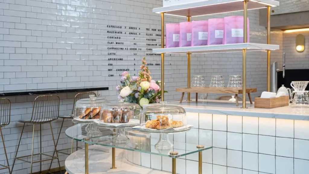 A modern cafe counter with glass displays of pastries, a flower arrangement, pink coffee bags on a shelf, and a white tile backdrop with a menu board.