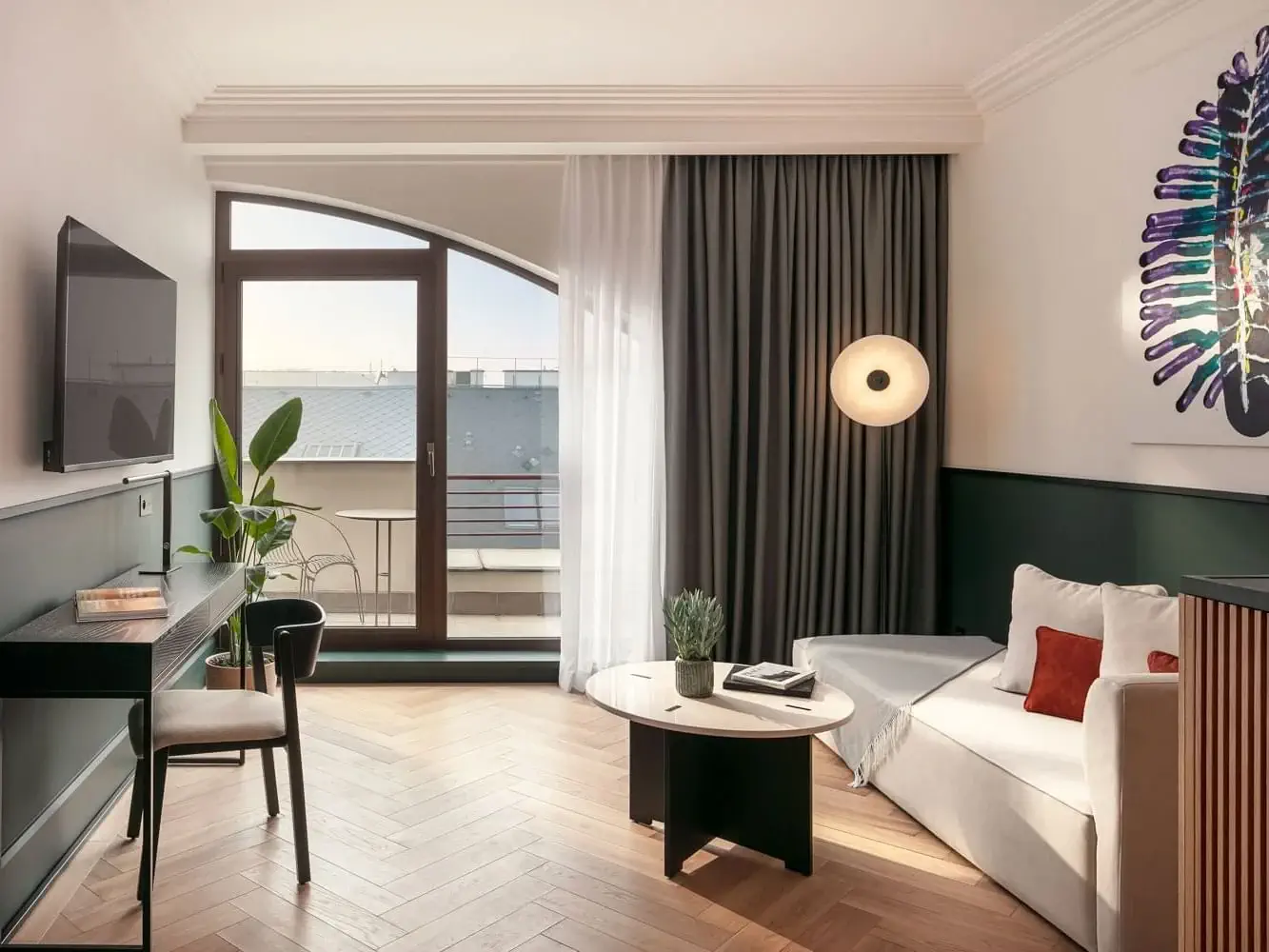 Světlý moderní obývací pokoj v Apartmá Executive s bílou pohovkou, kulatým konferenčním stolkem, psacím stolem s židlí, nástěnnou TV, velkým oknem se závěsy a balkonem.