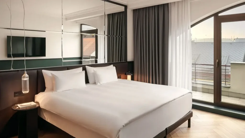 Eine moderne Executive Suite mit einem großen Bett mit weißer Bettwäsche, einem an der Wand montierten Fernseher, einer Spiegelwand, einer Hängelampe und einem Fenster mit Vorhängen, das zu einem privaten Balkon führt.