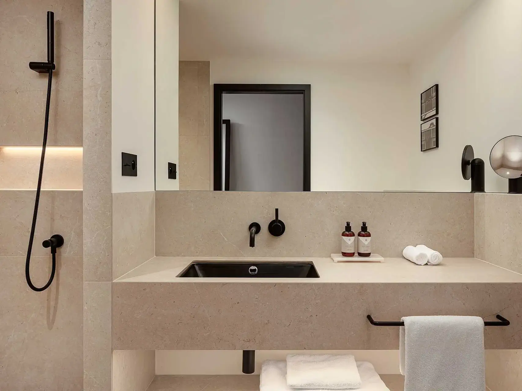 Modernes Deluxe-Zimmer-Badezimmer mit beigen Fliesen, schwarzen Armaturen, zwei Flaschen Seife, weißen Handtuchrollen und einer begehbaren Dusche auf der linken Seite.