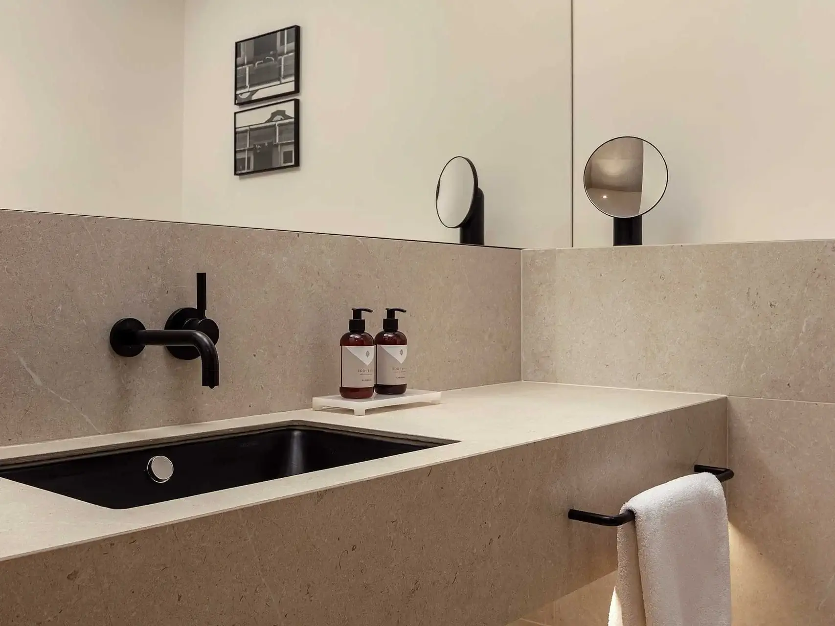 Modernes Deluxe-Zimmer-Badezimmer mit beiger Steinarbeitsplatte, schwarzem Waschbecken und Wasserhahn, zwei Seifenspendern, einem runden Spiegel, Handtüchern auf einer eleganten schwarzen Stange und gerahmten Fotos an der Wand.