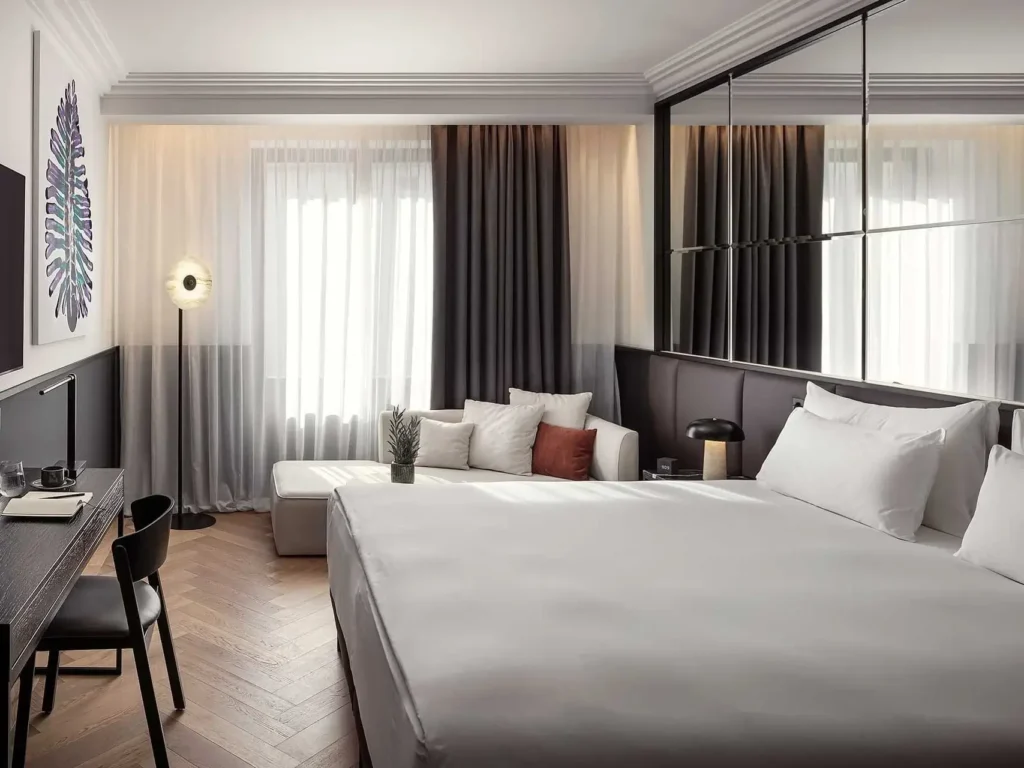 Modernes Deluxe-Zimmer mit großem Bett, Schreibtisch mit Stuhl, Sofa am Fenster, Stehlampe, Wandkunst und neutralen Farbtönen.